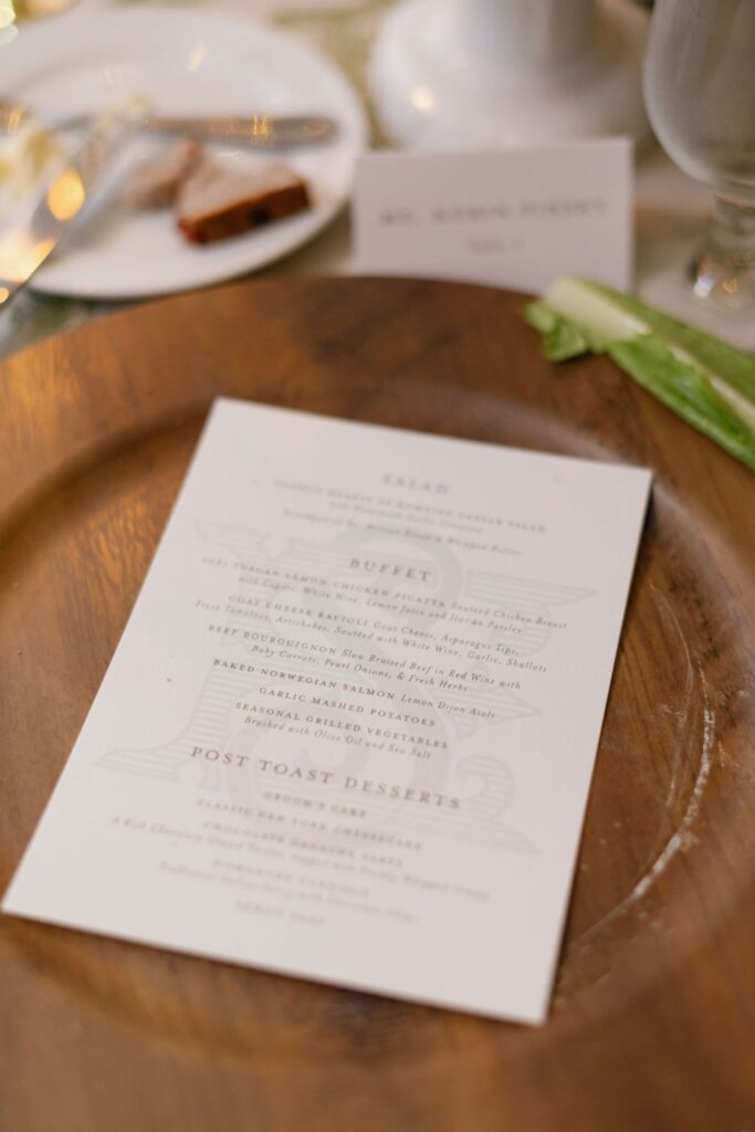 A close-up of a wedding dinner menu at a Chastain Horse Park rehearsal dinner, placed on a wooden charger plate. The blurred background shows part of a table setting with food, a wine glass, and a partially readable name card.