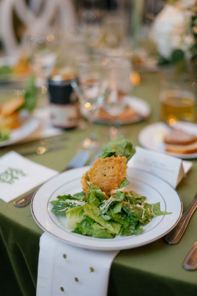 A plate of fresh green salad topped with a crispy cheese wafer is set on a green tablecloth at an elegant table setting—perfect for a Chastain Horse Park rehearsal dinner.