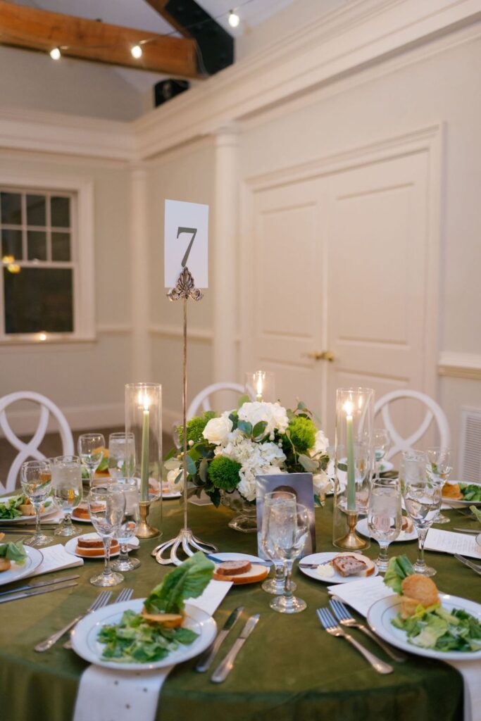 A round table set for a formal Chastain Horse Park rehearsal dinner features a green tablecloth, white plates with salad, glasses, bread, candles, a white floral centerpiece, and table number 7. White chairs surround the elegant setting.