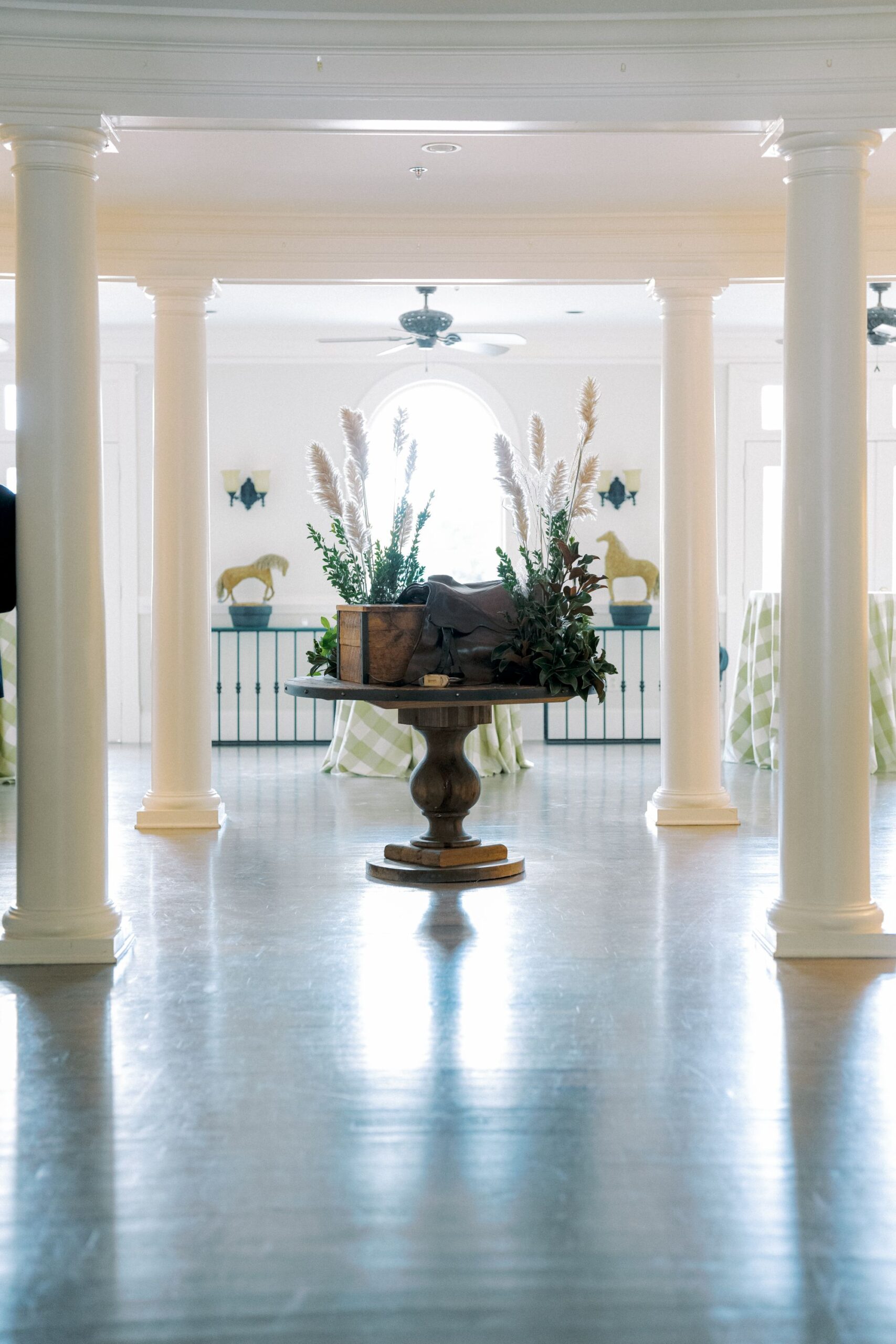 A round wooden table with plants and decorative items stands between white columns in a bright, airy room with shiny floors and light streaming in from large windows.