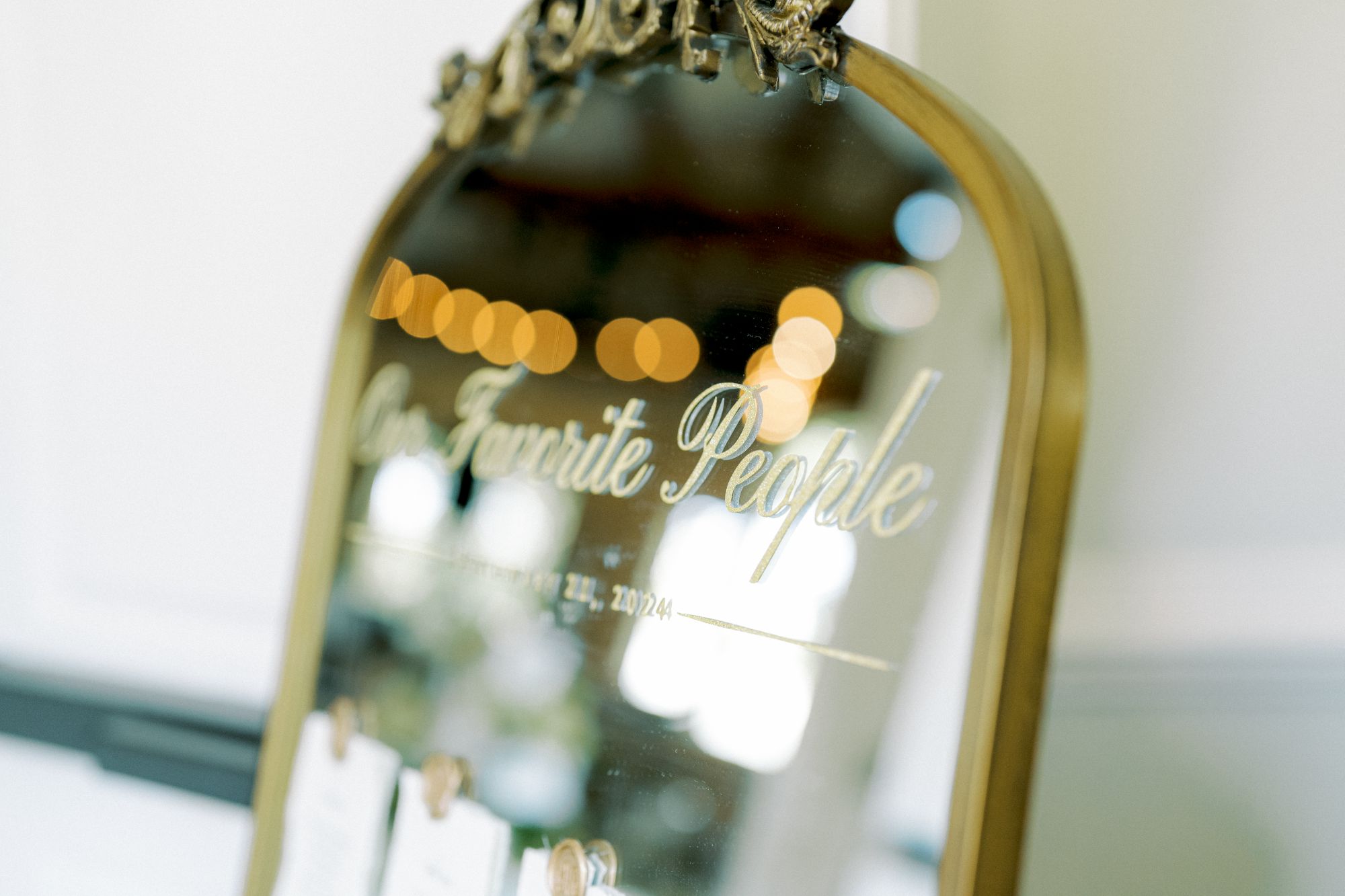 A gold-framed mirror with the words “Our Favorite People” written on it in elegant script, blurred lights reflected at the top and paper cards or notes attached at the bottom.