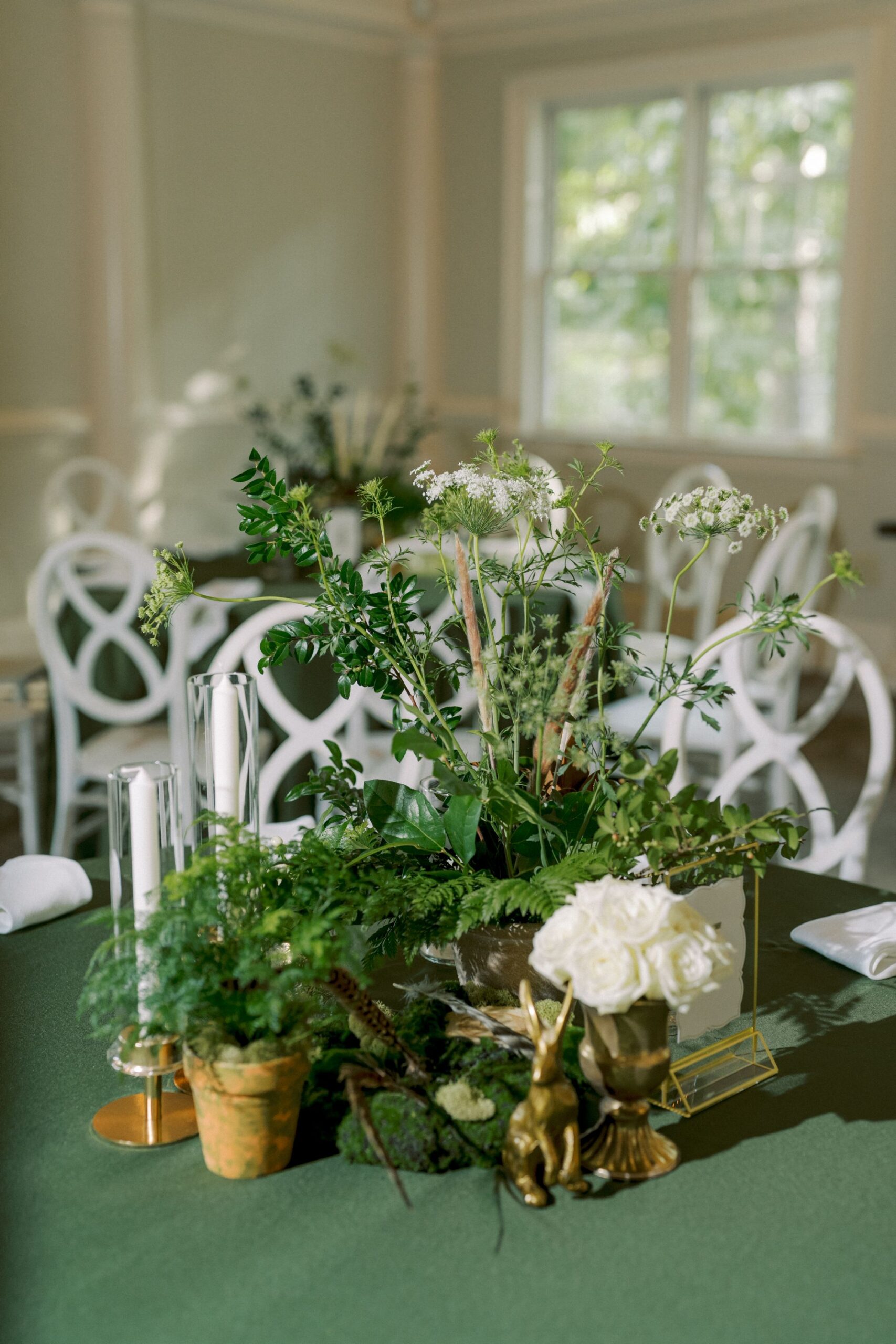 A table with a green tablecloth decorated with potted plants, white flowers, two tall candles, and a small gold rabbit figurine. White chairs and a window are visible in the softly lit background.