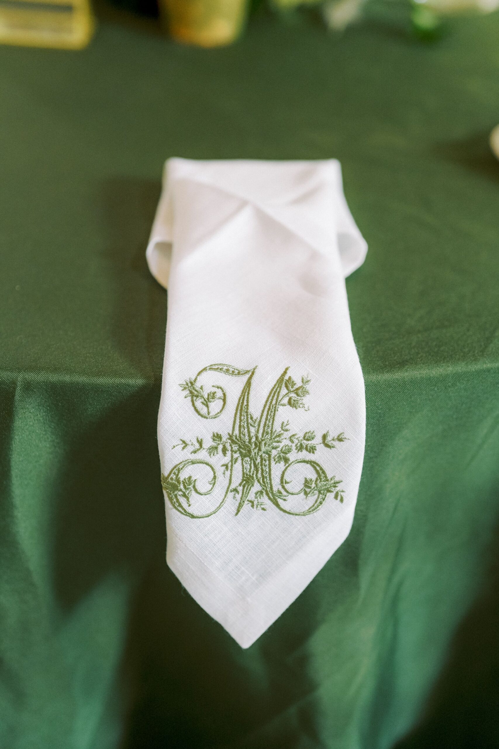 A white napkin with a green embroidered letter M and decorative floral designs is neatly folded and placed on a green tablecloth.