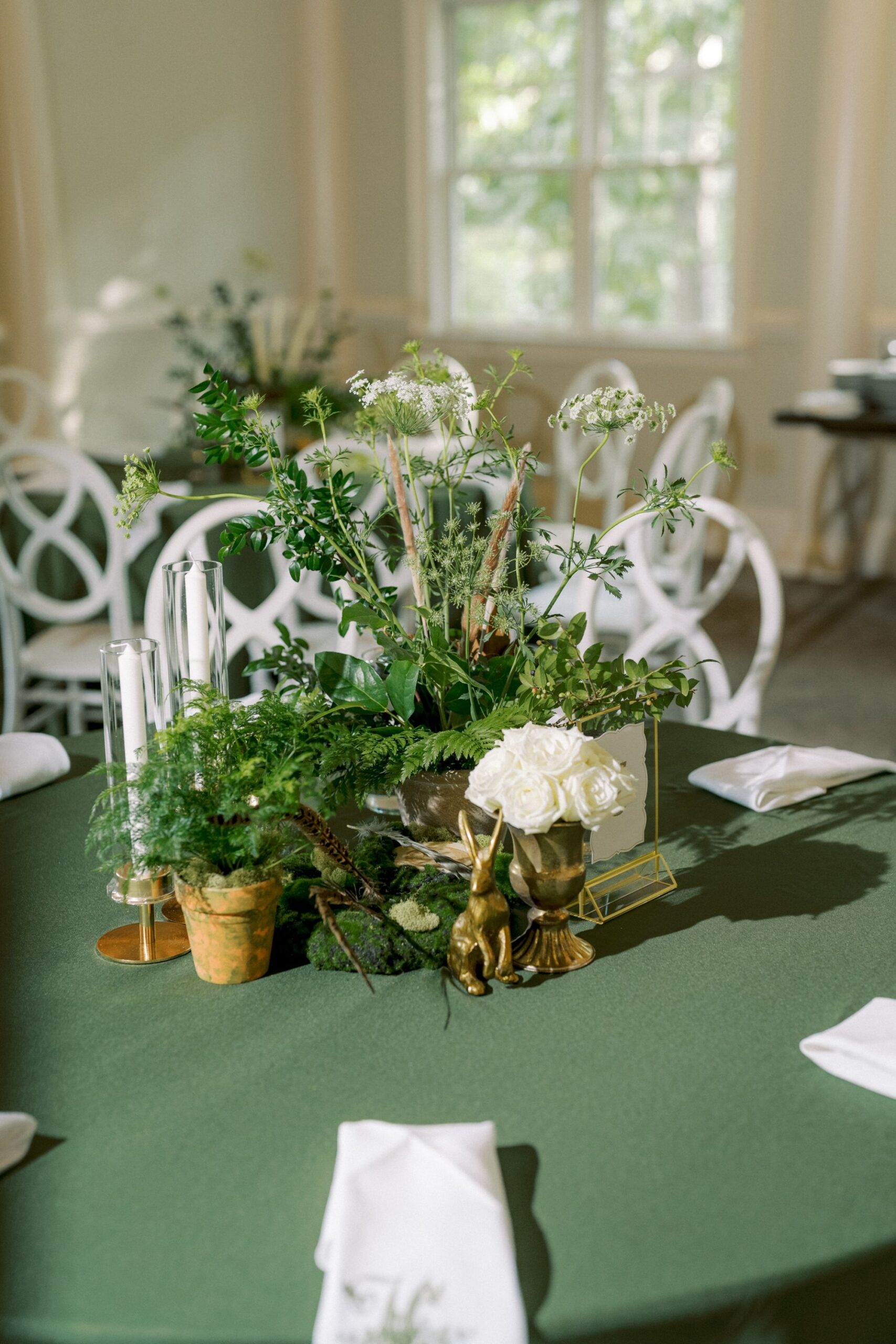 A green tablecloth with white napkins, a centerpiece featuring lush greenery, white roses, candles, a small potted plant, and a gold rabbit figurine, set in a sunlit room with white chairs and large windows.