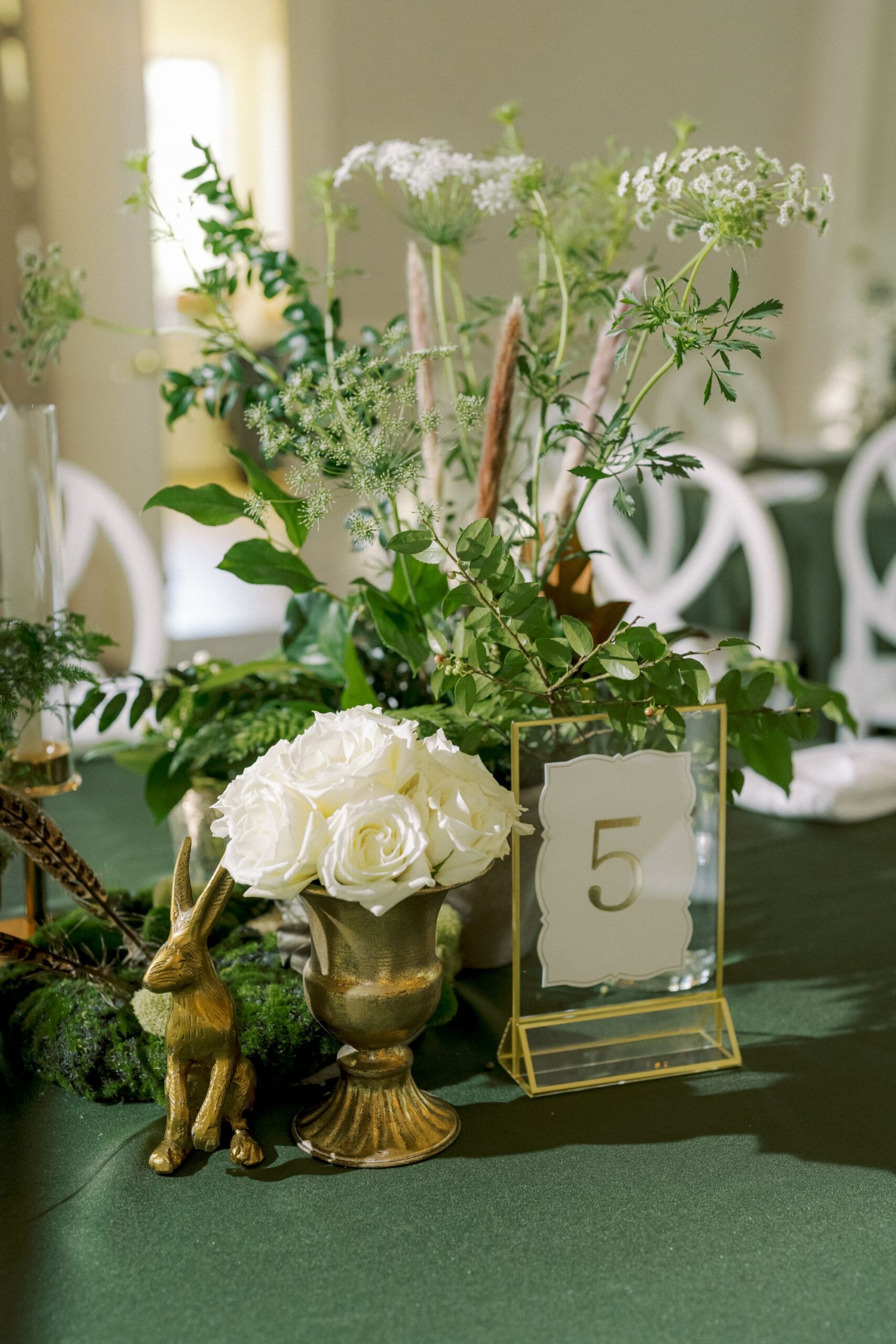 A table centerpiece features a gold vase with white roses, lush greenery, a gold rabbit figurine, and a clear table number sign with the number 5. The background shows white chairs and a green tablecloth.