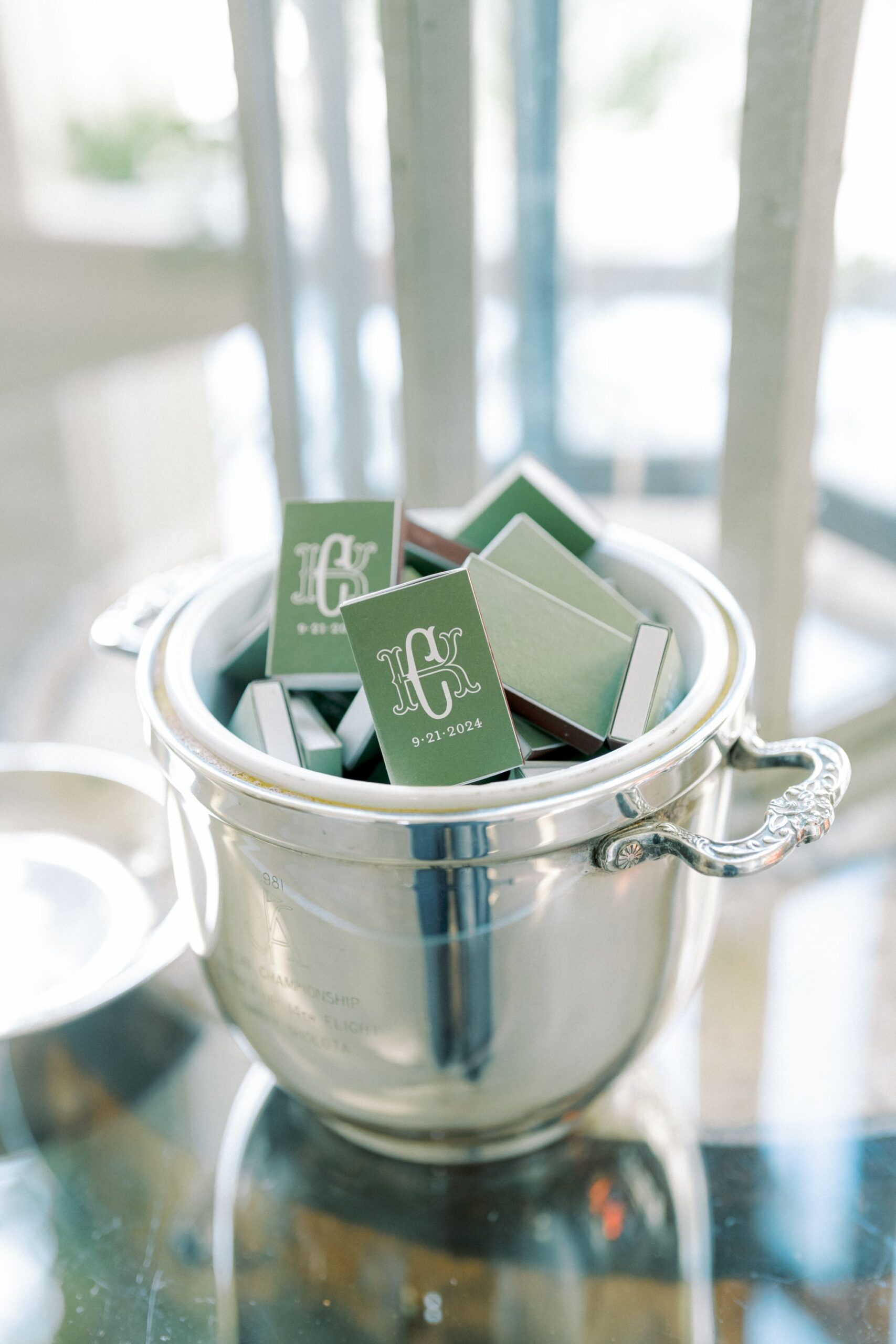 A silver ice bucket filled with green matchboxes featuring a white monogram and date, sitting on a reflective glass surface.
