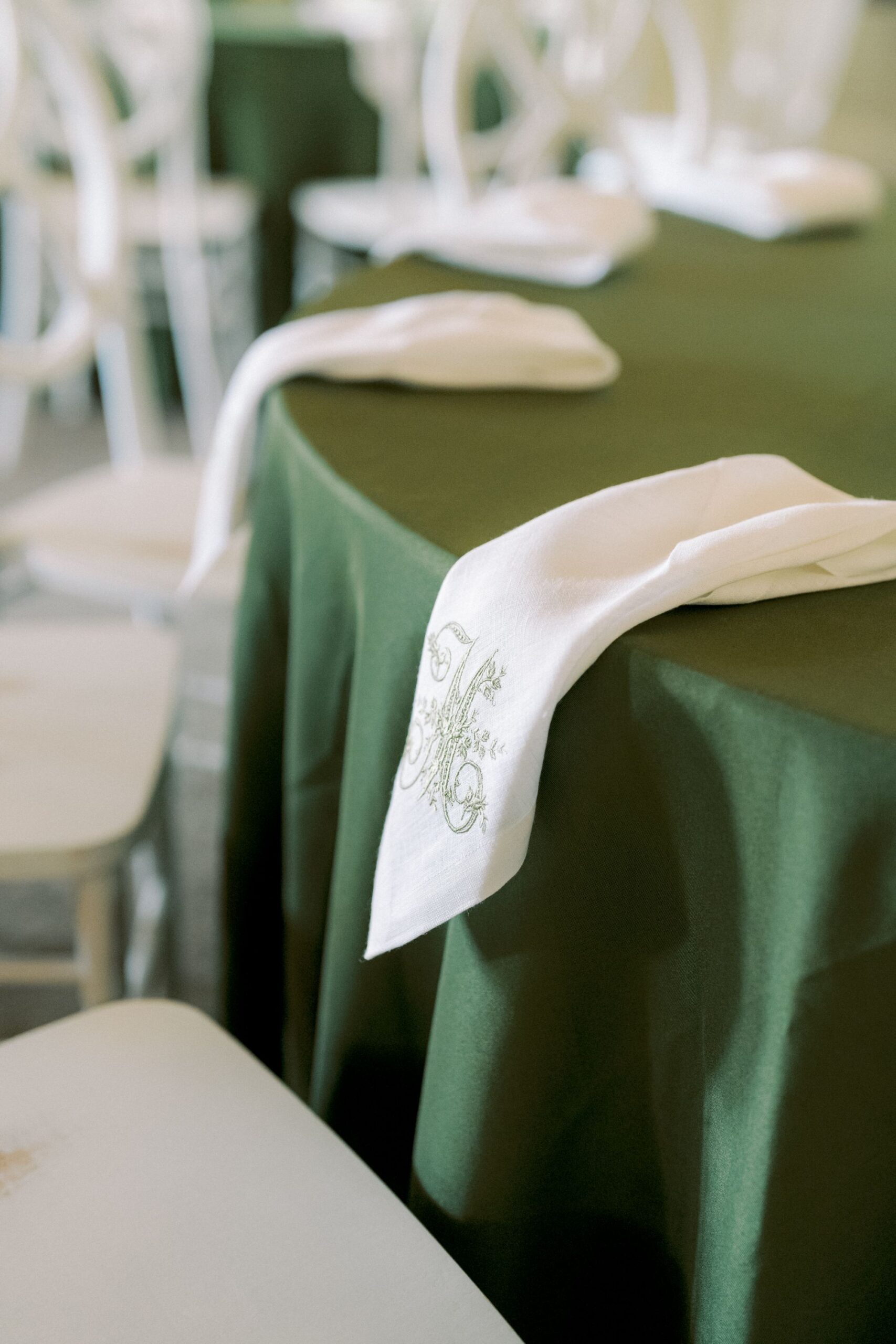 A close-up of a round table covered with a dark green tablecloth, featuring a white napkin embroidered with a decorative letter placed on the edge. White chairs surround the table.