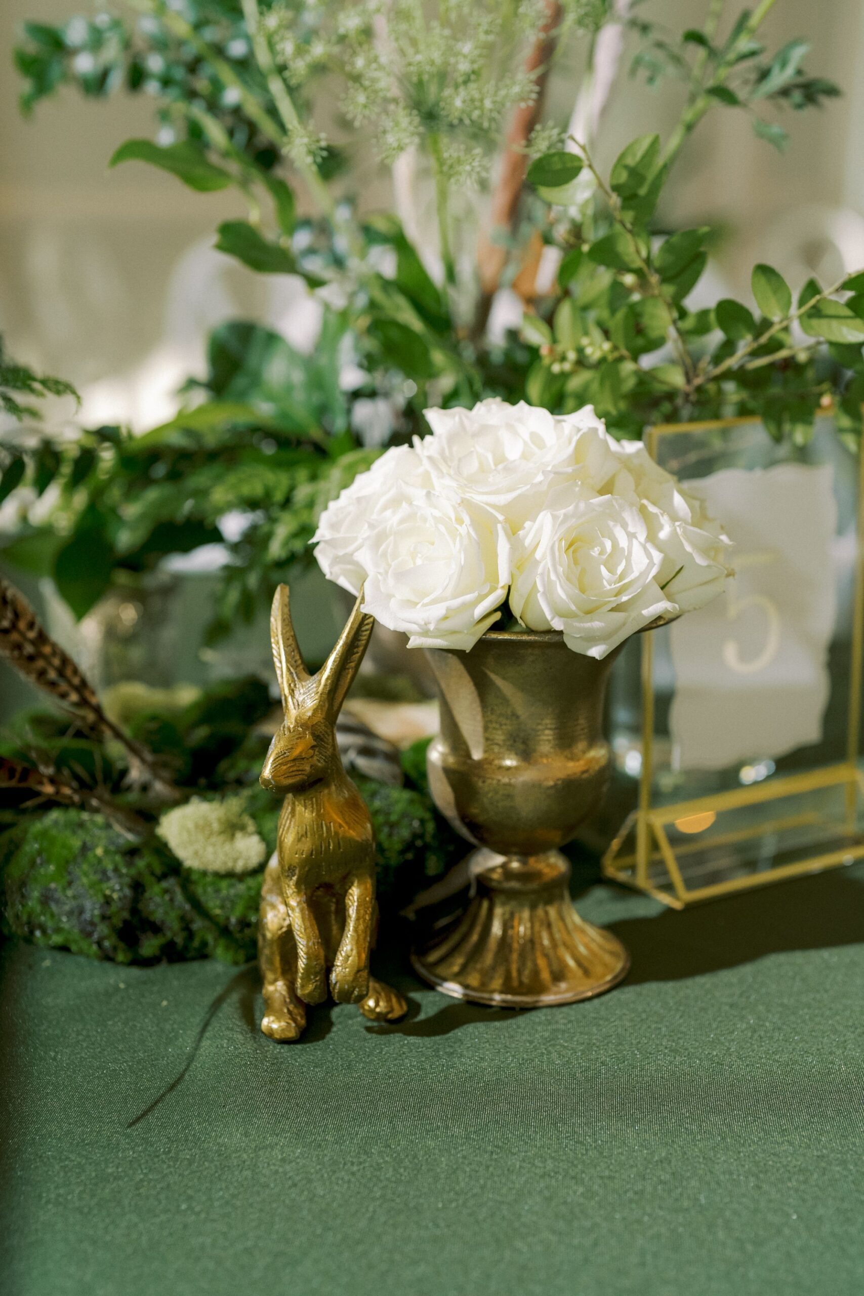 A gold vase with white roses sits next to a small gold rabbit statue on a green tablecloth, surrounded by greenery and moss, creating an elegant, natural-themed centerpiece.