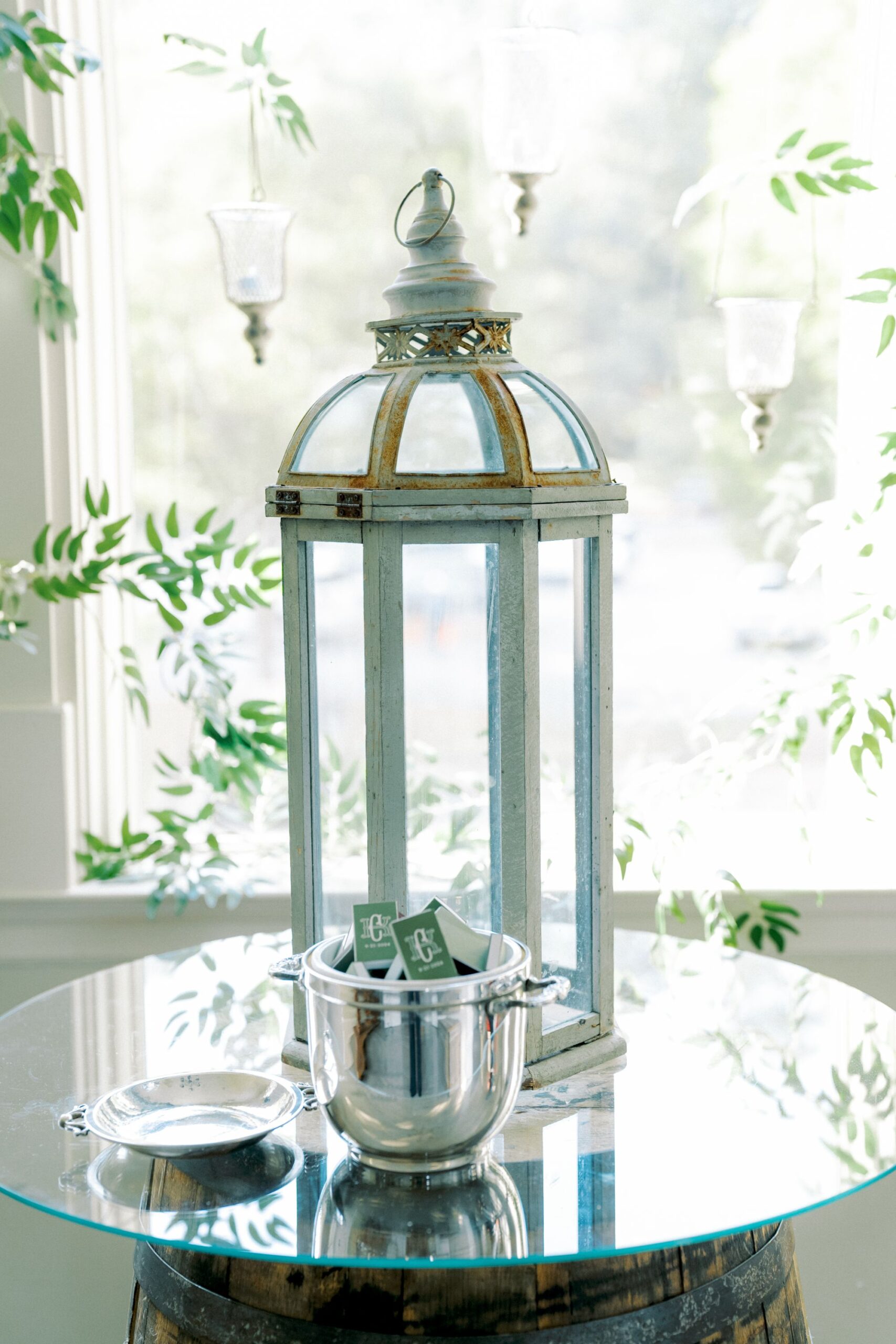 A tall, decorative lantern sits on a glass-topped barrel. In front of it, a silver bowl holds green and white sugar packets, with a lid resting beside it. Lush greenery and hanging glass votives are in the bright background.