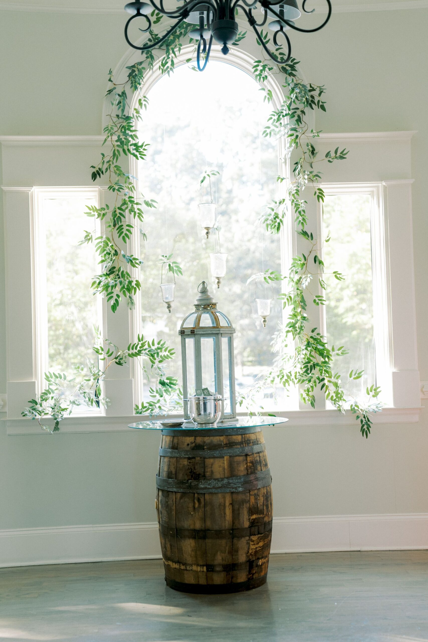 A rustic wooden barrel topped with a glass surface holds a decorative lantern and ice bucket, set in front of a large arched window with greenery vines draped around it. Soft natural light fills the scene.