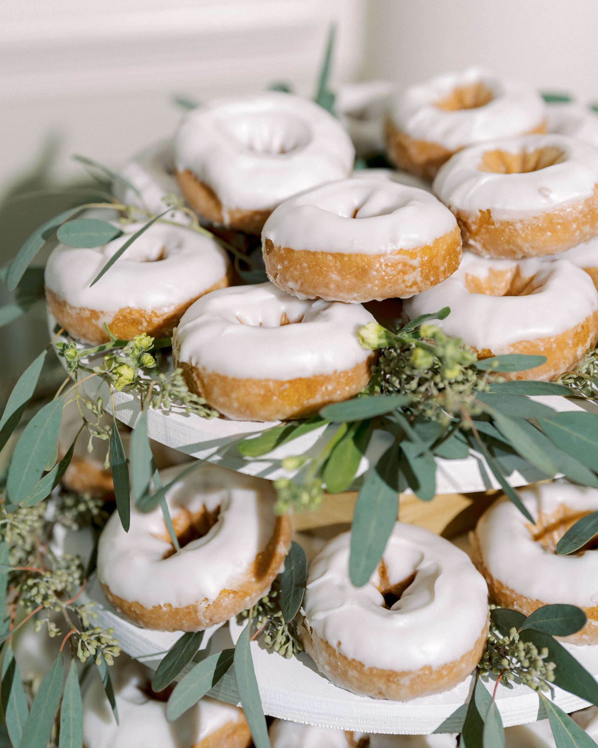 A display of glazed donuts stacked on a tiered stand, decorated with green leaves and small sprigs of flowers.
