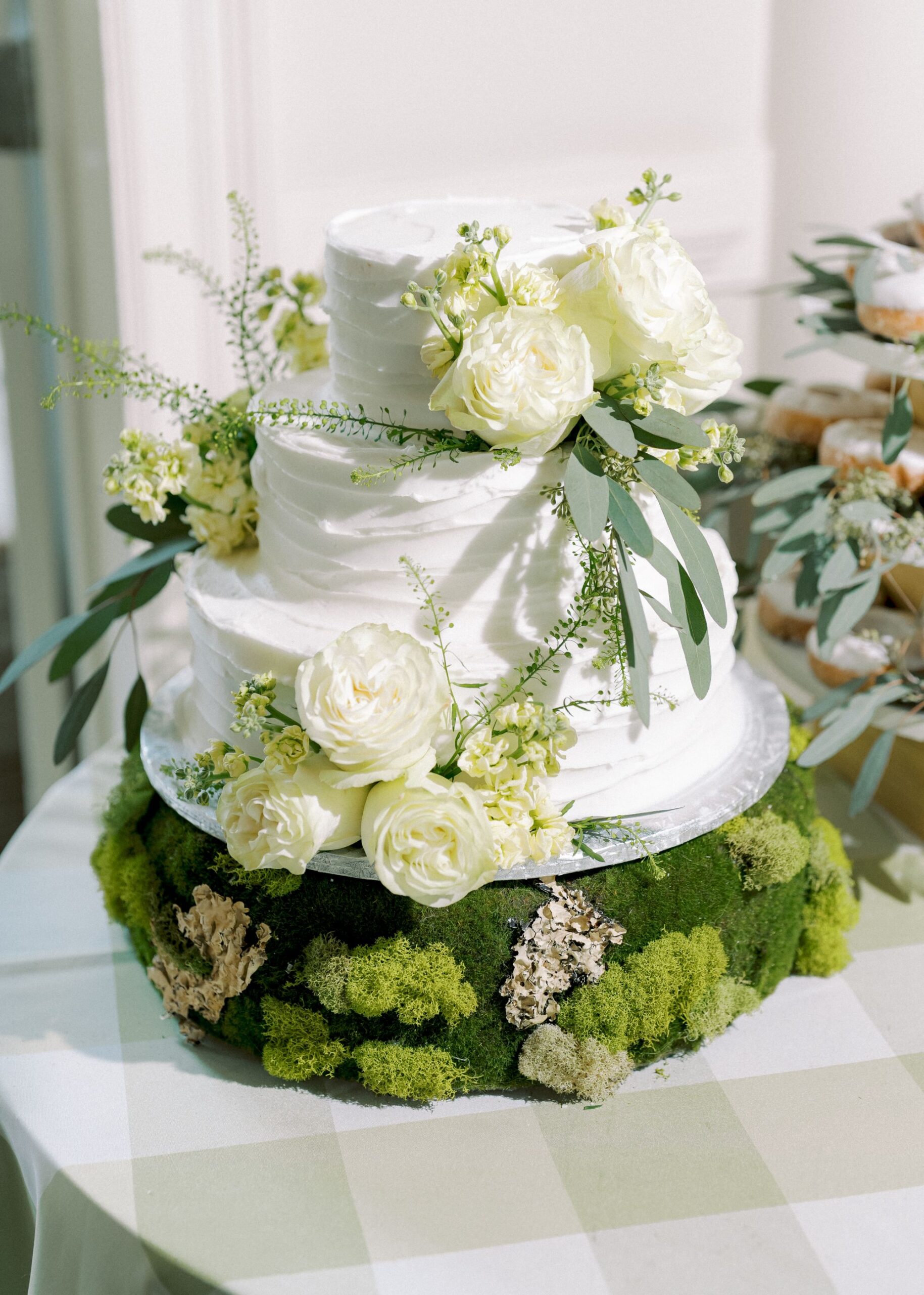 A two-tiered white cake decorated with white roses and greenery sits on a base covered in green moss, displayed on a checkered tablecloth.