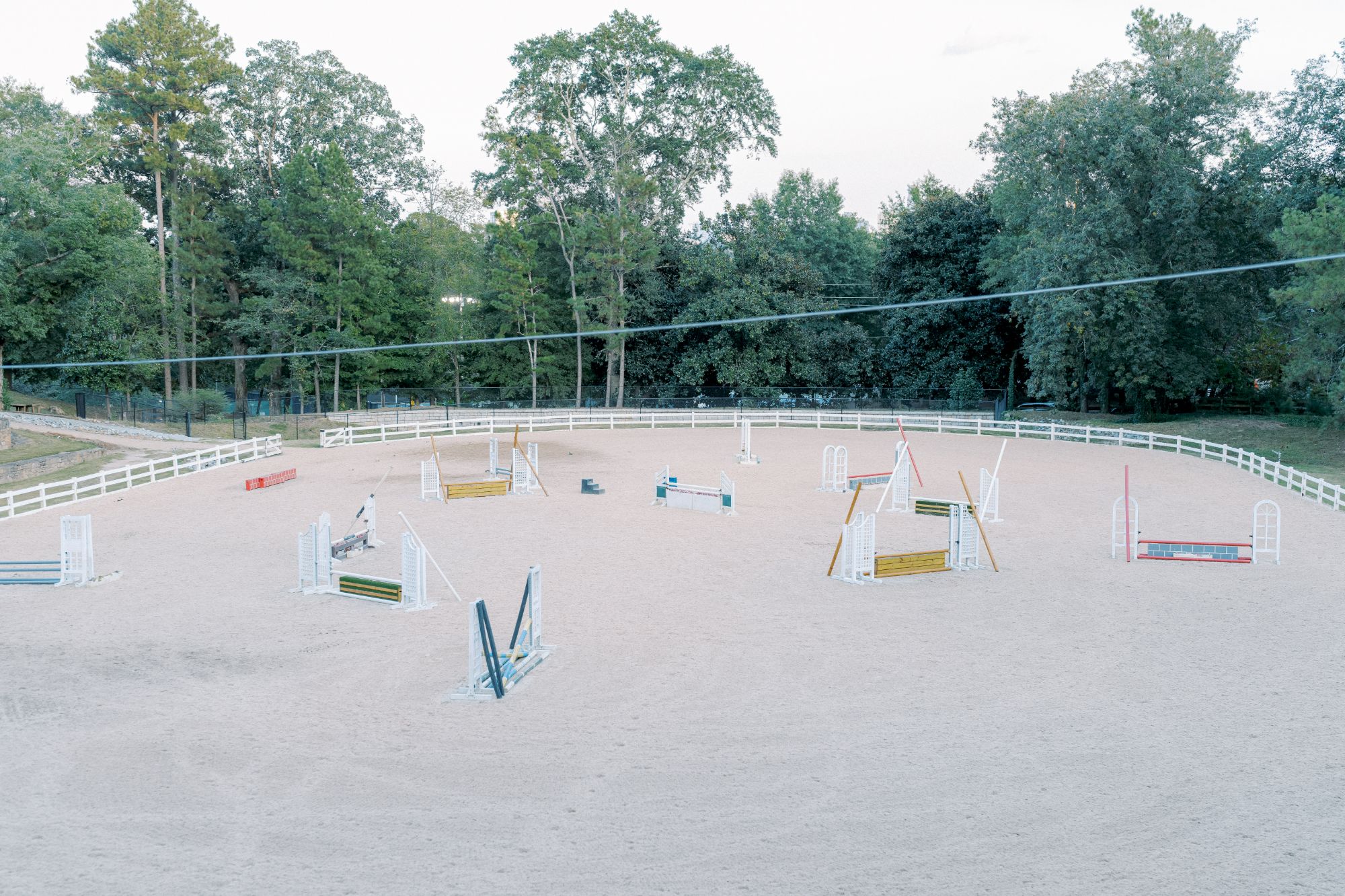 An outdoor equestrian arena with sandy ground containing several colorful horse jump obstacles, surrounded by a white fence and green trees in the background.