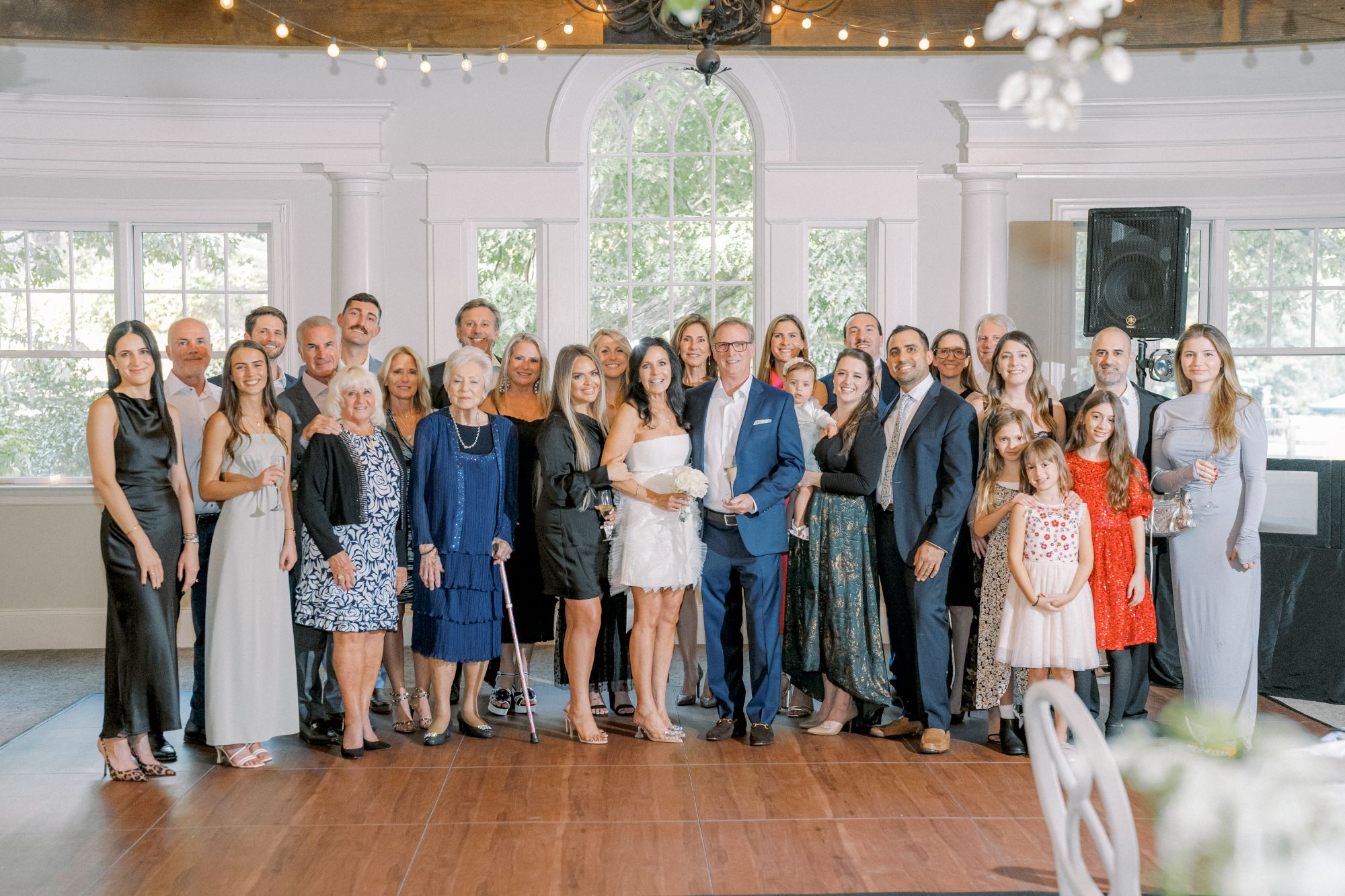 A large group of people, dressed in semi-formal attire, pose together indoors in front of tall windows. Some are smiling, and a couple stands at the center holding hands. Warm lighting creates a festive atmosphere.