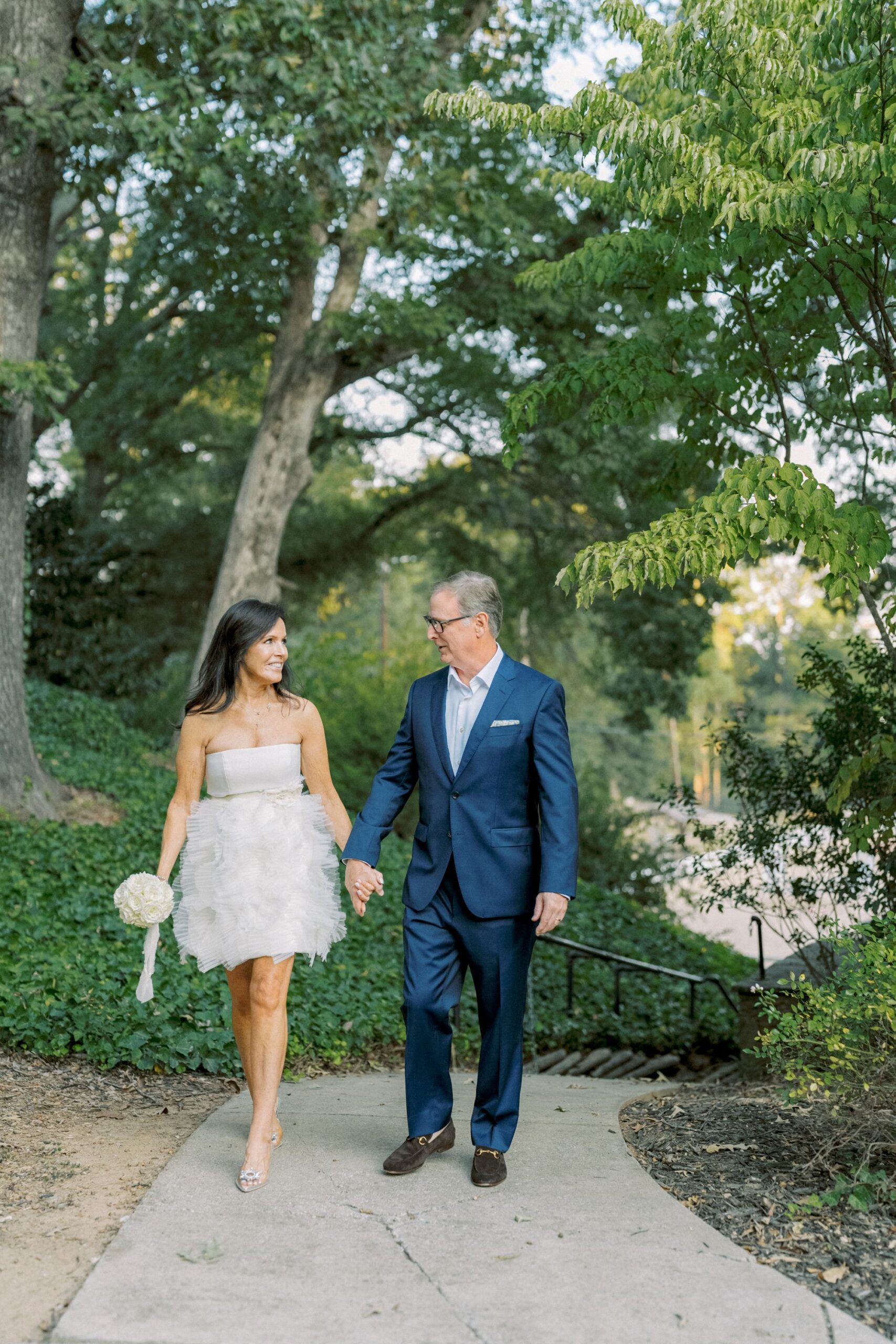 A couple walks hand in hand outdoors; the woman wears a short white dress and holds a bouquet, while the man wears a dark suit. They smile at each other on a tree-lined path.