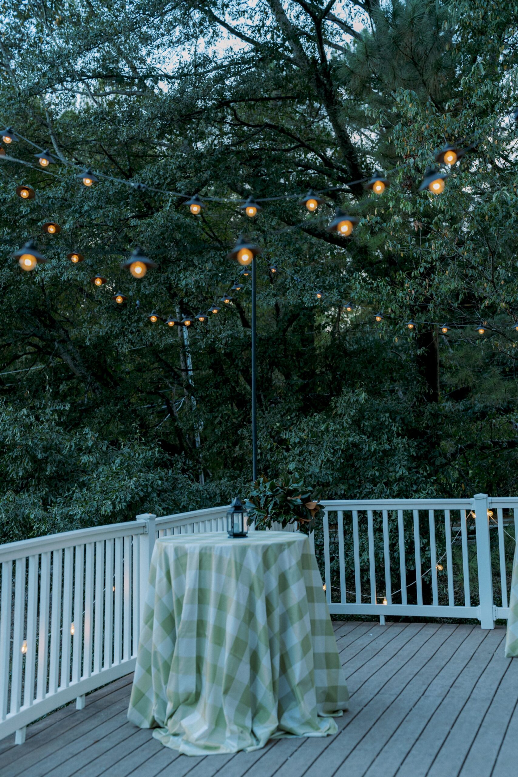 A table covered with a green and white checkered cloth stands on a wooden deck with a white railing, surrounded by trees and string lights glowing overhead in the evening.