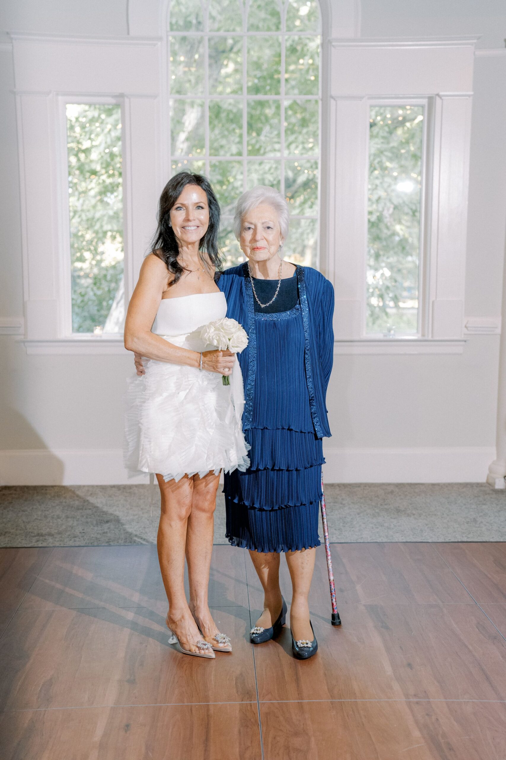 Two women stand side by side indoors. One wears a short white dress and holds a bouquet; the other wears a blue dress and holds a cane. They smile in front of tall windows with trees visible outside.