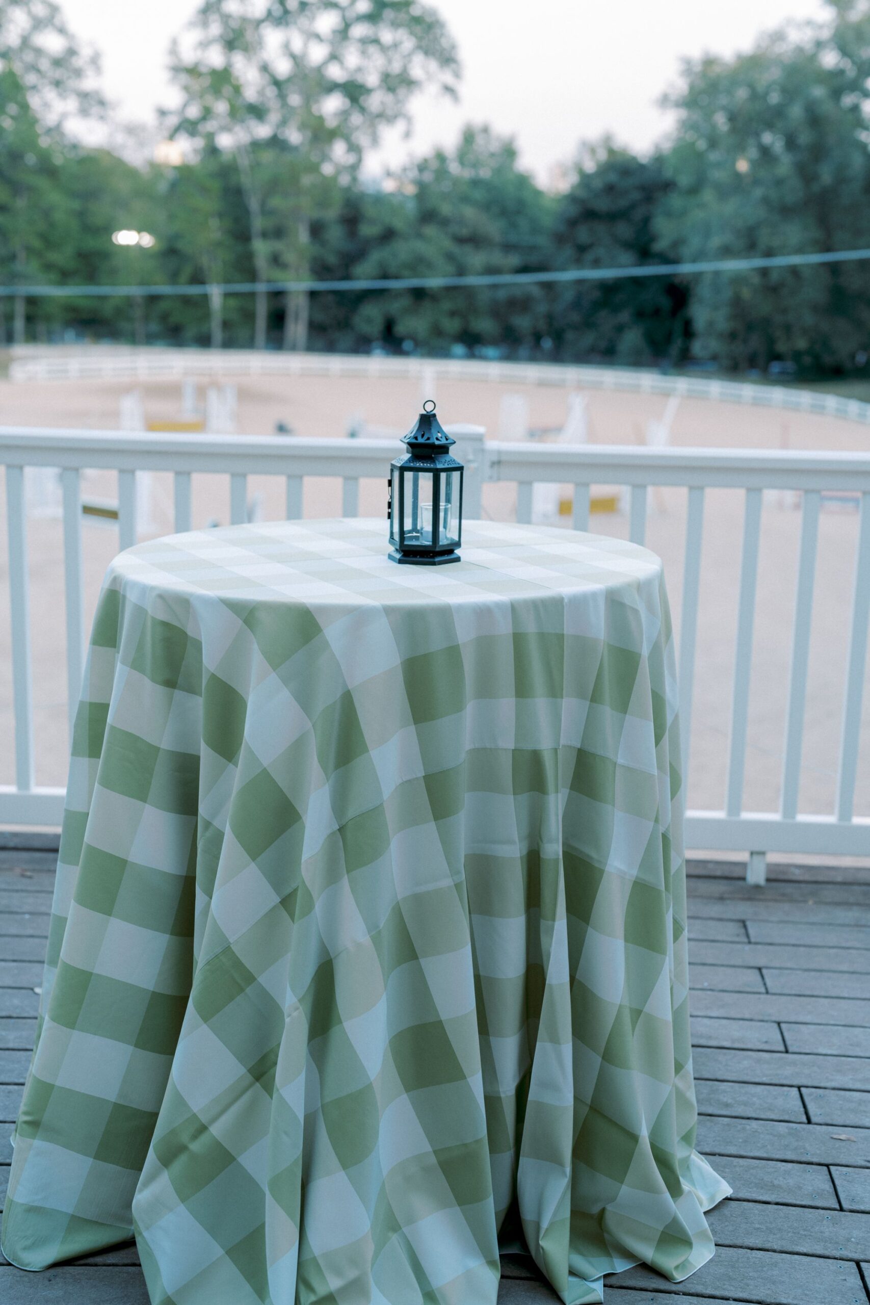 A high-top table covered with a green and white checkered tablecloth sits on a wooden deck, topped with a small decorative lantern. A white railing and blurry trees are visible in the background.