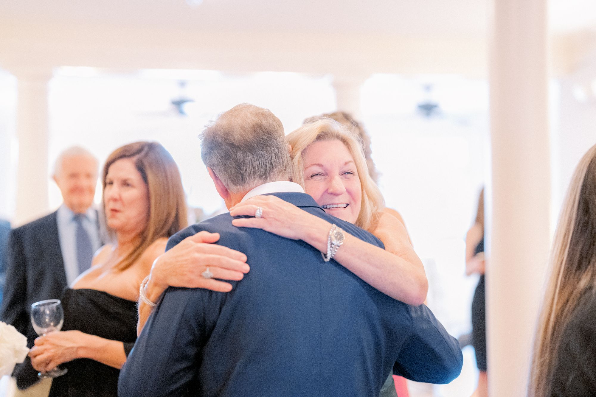 A smiling woman hugs a man in a suit at a bright indoor gathering. Other people, dressed formally, are visible in the softly blurred background.