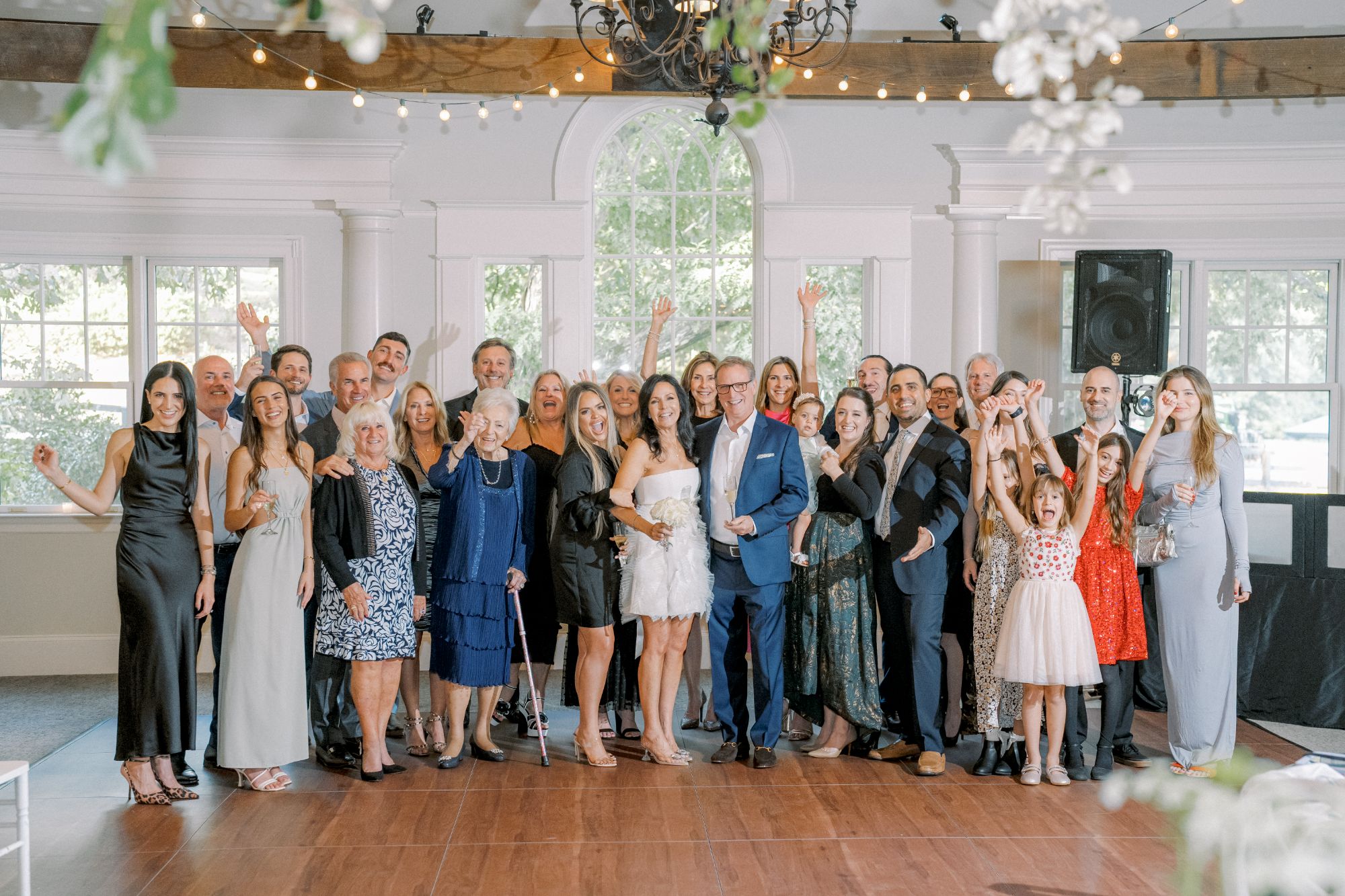 A large group of people of various ages dressed in semi-formal attire poses together indoors, smiling and waving at the camera in front of a bright window and chandelier.