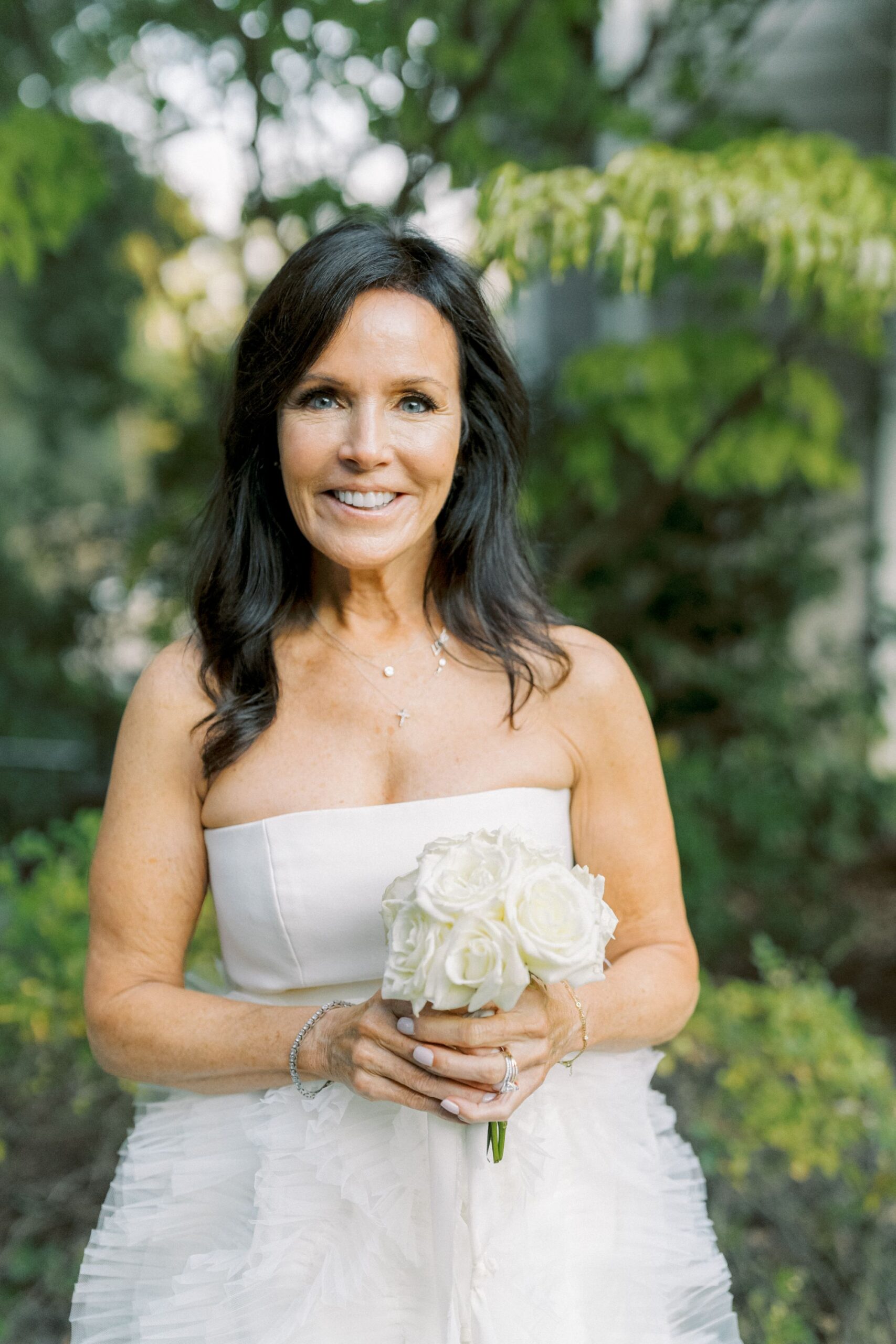 A woman with long dark hair wearing a strapless white dress holds a bouquet of white roses and smiles, standing outdoors with green foliage in the background.