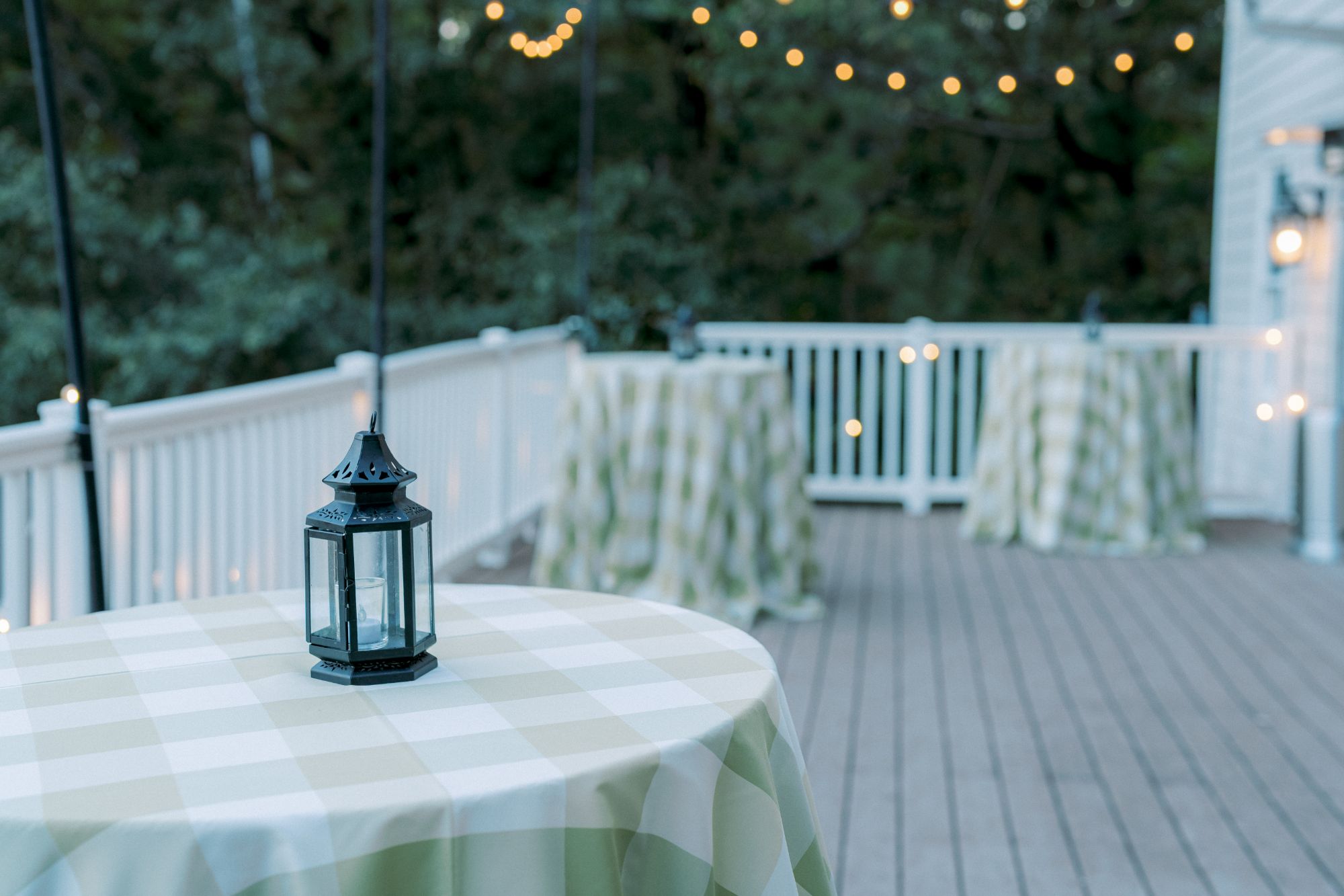 A small lantern sits on a checkered table covered in a green and white cloth on an outdoor patio with string lights and trees in the background.