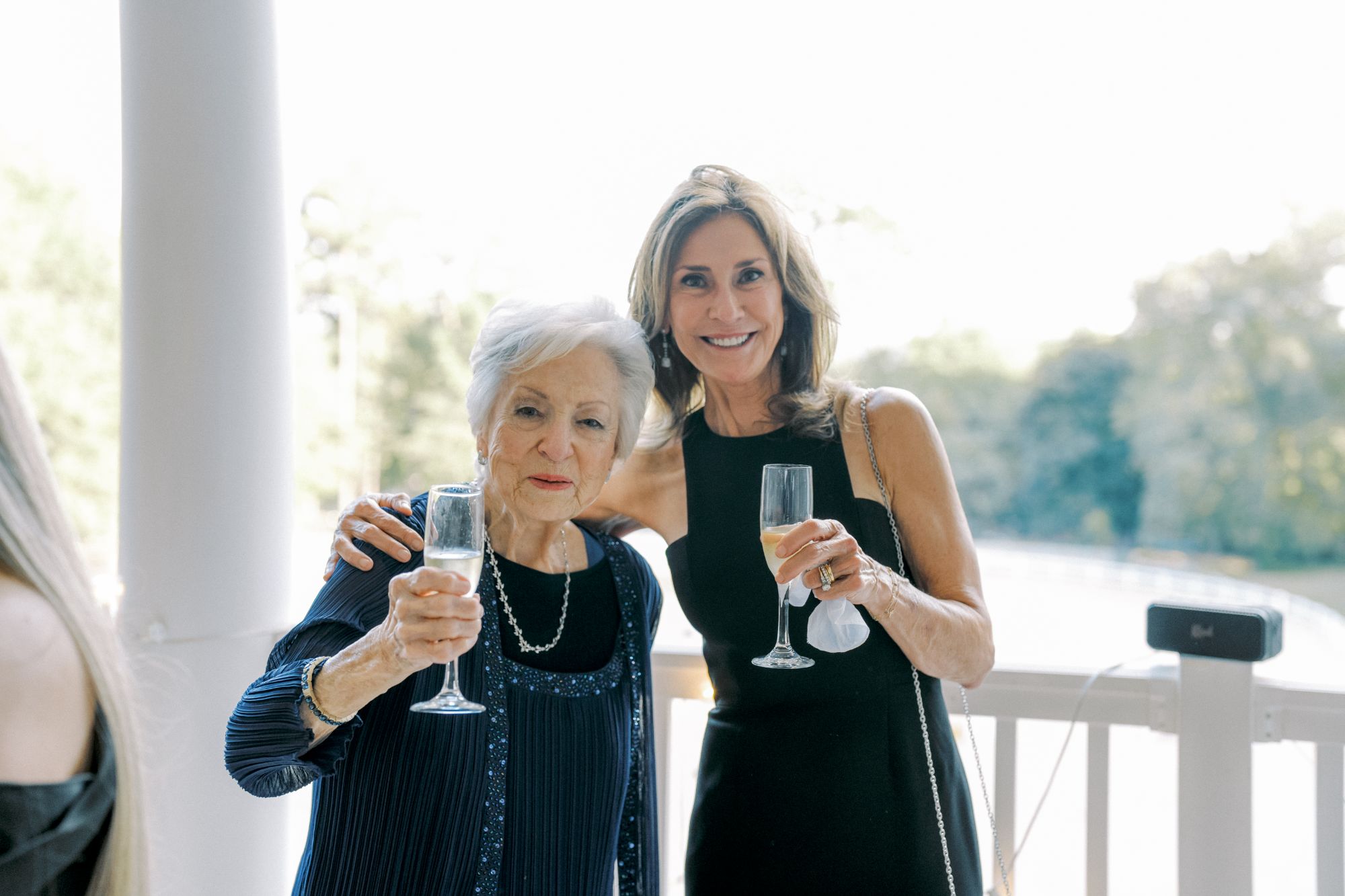 Two women dressed formally stand close together, smiling and holding champagne glasses on a porch with trees in the background. One woman has gray hair and wears a blue outfit; the other has light brown hair and wears black.
