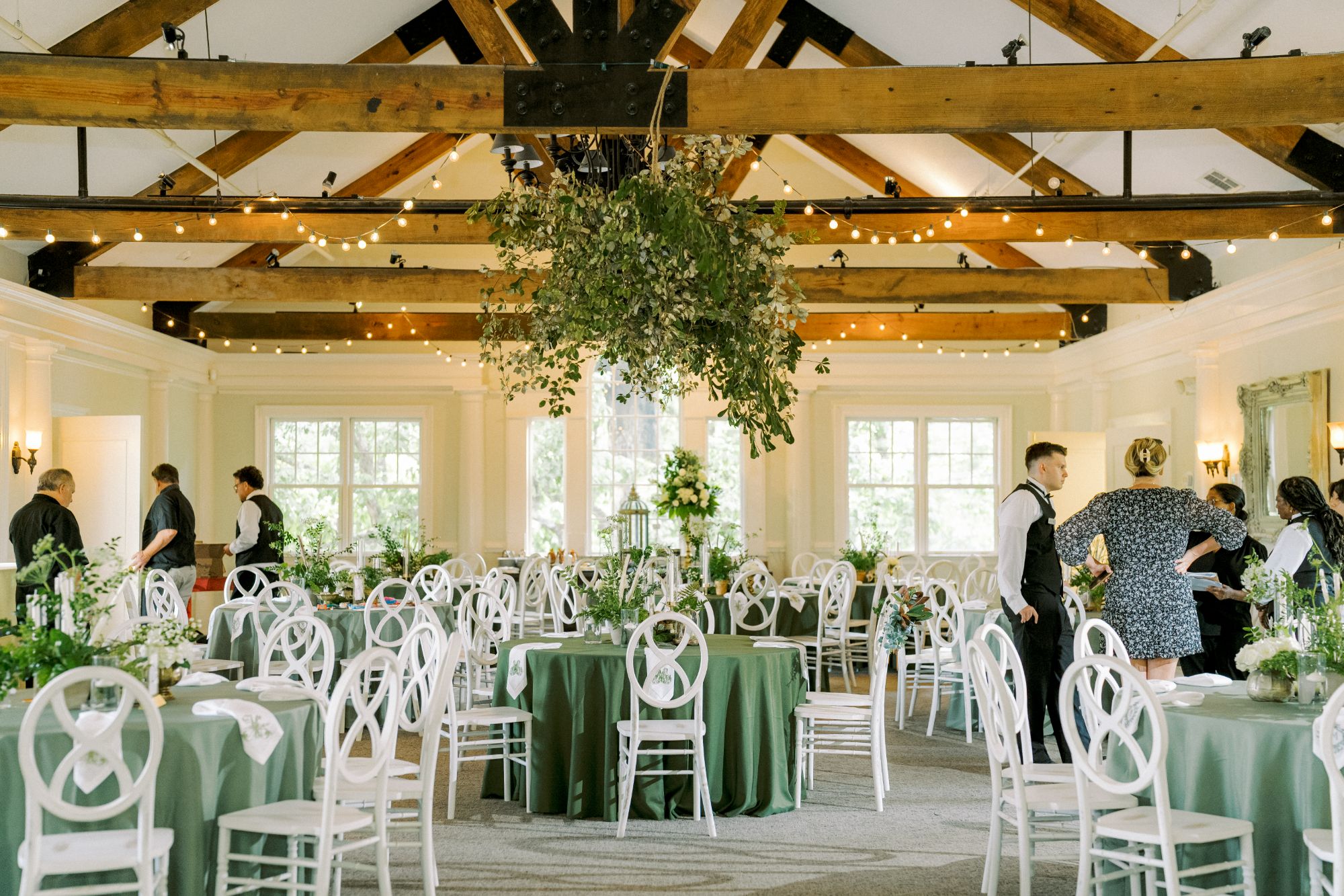 Elegant banquet hall with wooden beams, green tablecloths, white chairs, and lush greenery centerpieces. Waitstaff in black vests prepare tables, and string lights hang overhead, creating a warm, inviting atmosphere.
