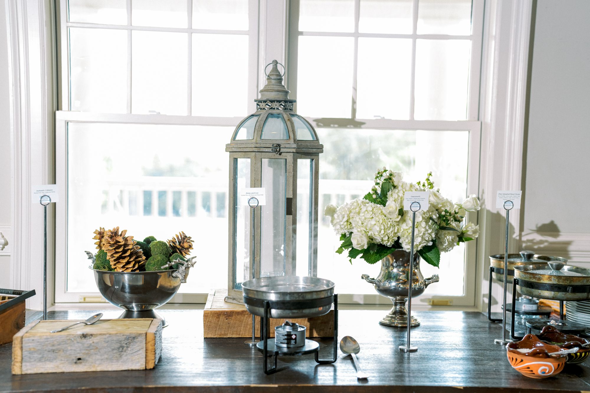 A buffet table with a large decorative lantern, a bowl of pinecones, a bouquet of white flowers, and several serving dishes, set in front of bright windows.