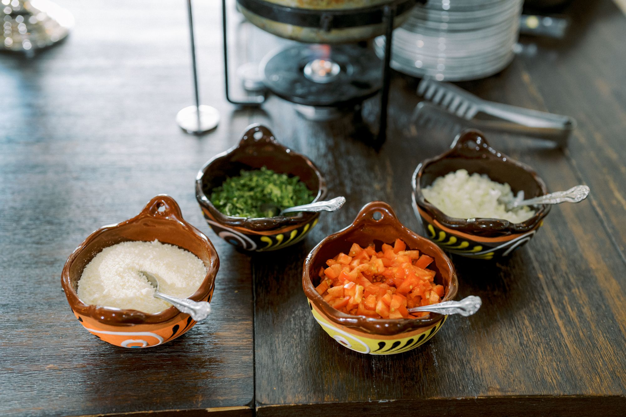 Four small brown bowls with silver spoons, each containing different chopped ingredients: sugar, green herbs, diced red bell pepper, and chopped onions, arranged on a wooden table.