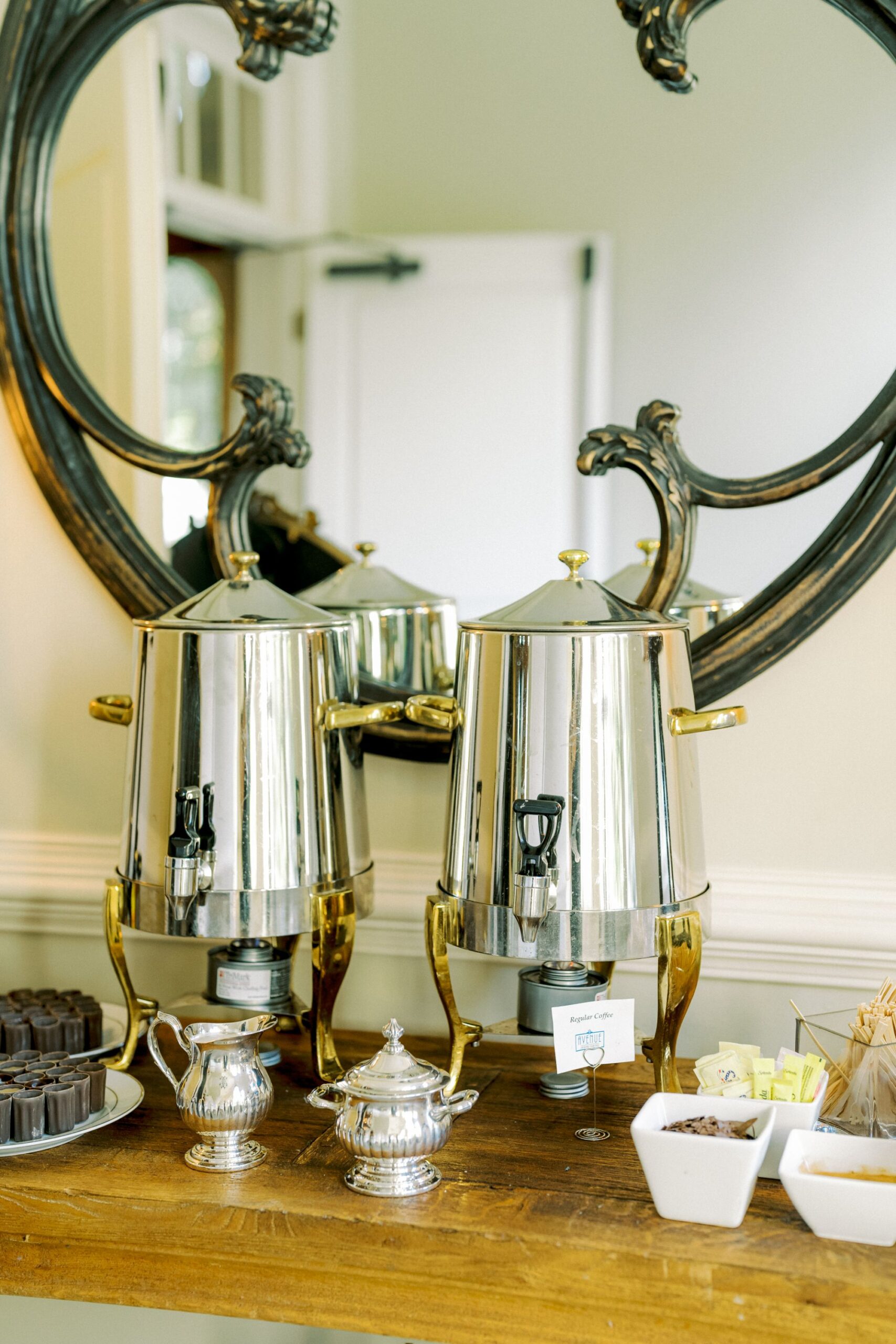 Two shiny metal beverage dispensers with spigots sit on a wooden table in front of a decorative mirror, surrounded by a sugar bowl, cream pitcher, sweetener packets, and cups of chocolate and sugar cubes.