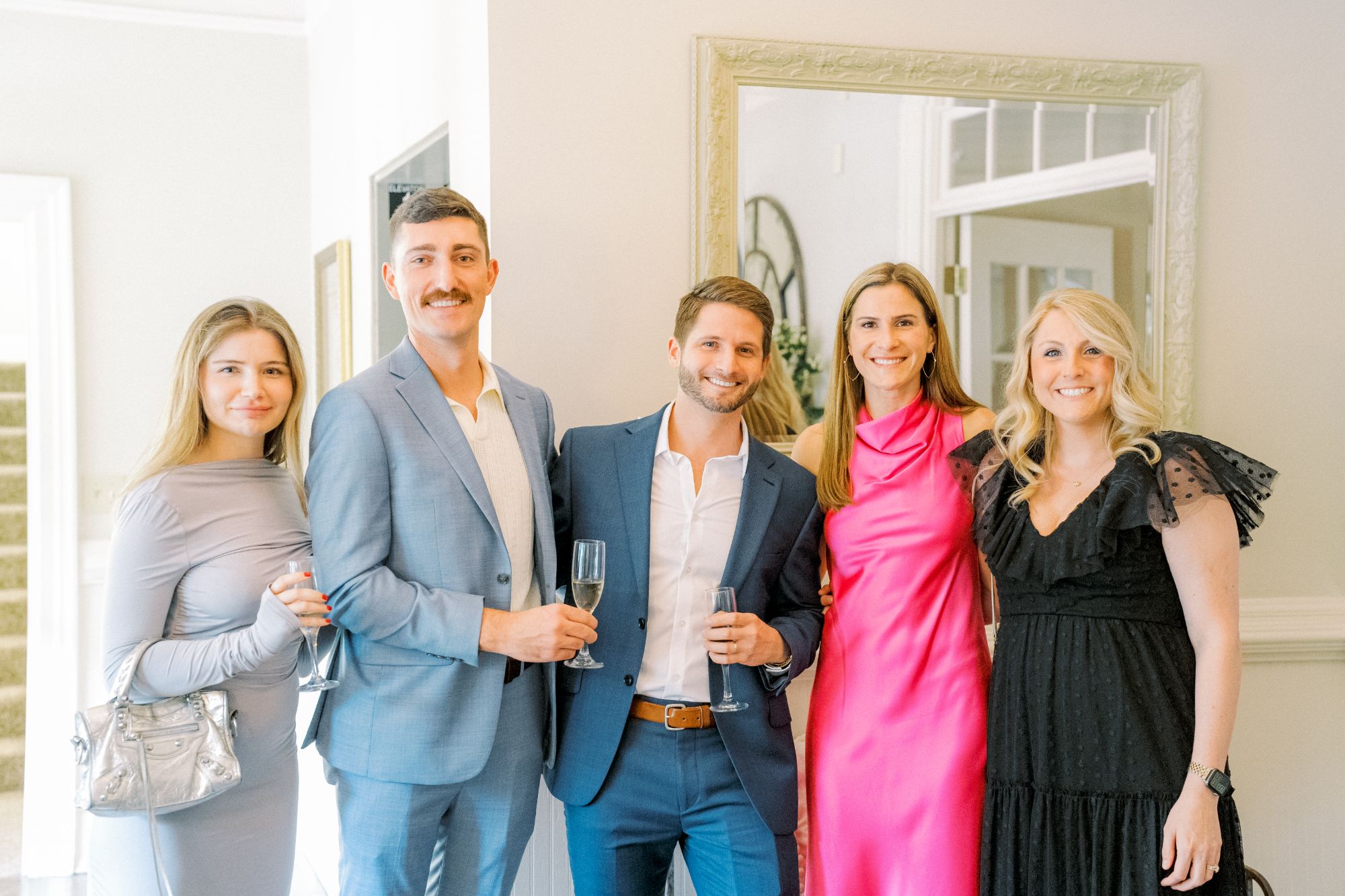 Five people dressed in semi-formal attire stand indoors, smiling at the camera and holding drinks. There is a mirror and white walls in the background, with daylight coming through nearby windows.