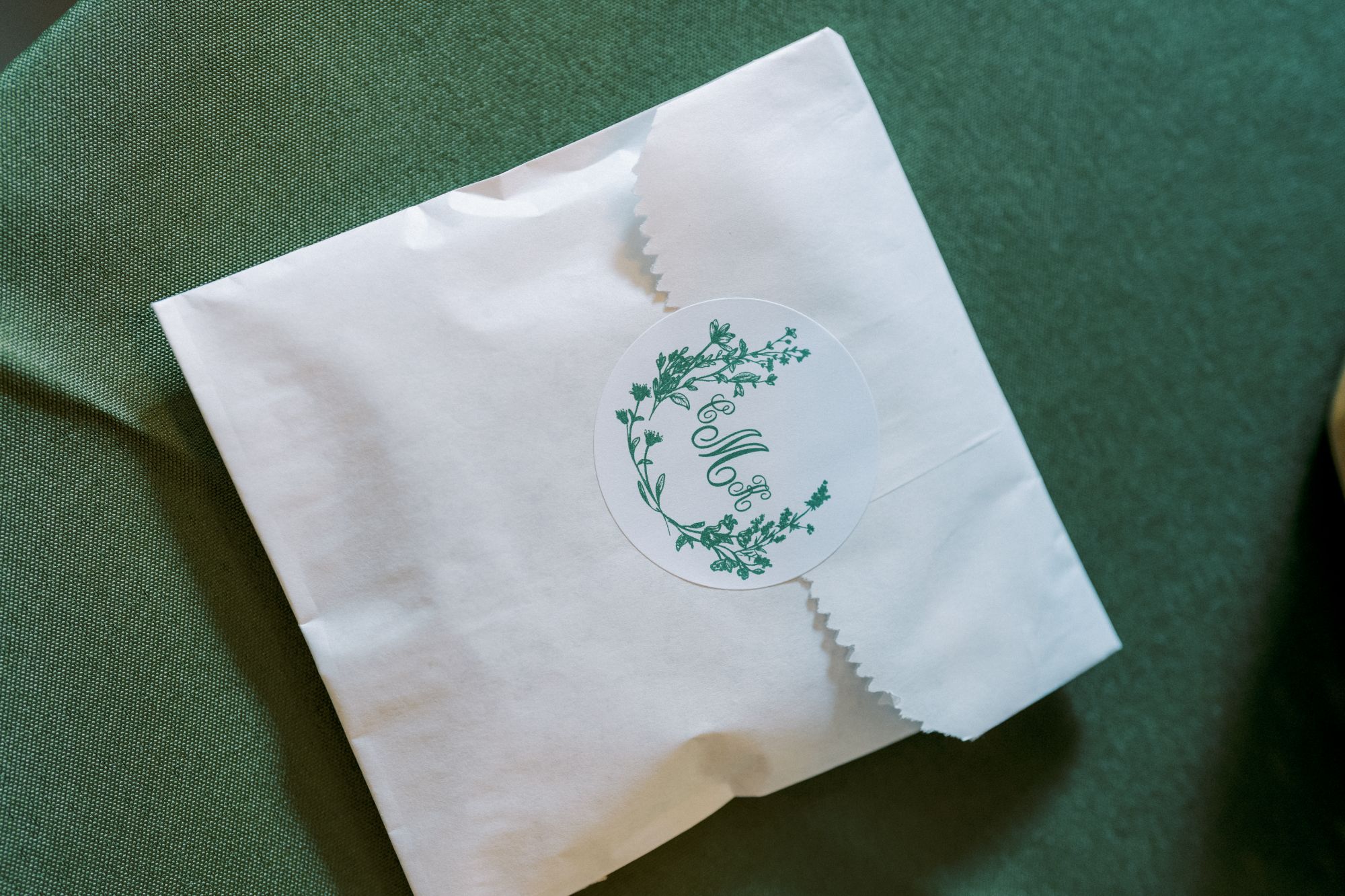 A white paper bag sealed with a round sticker featuring green floral designs and elegant script initials, placed on a green textured surface.