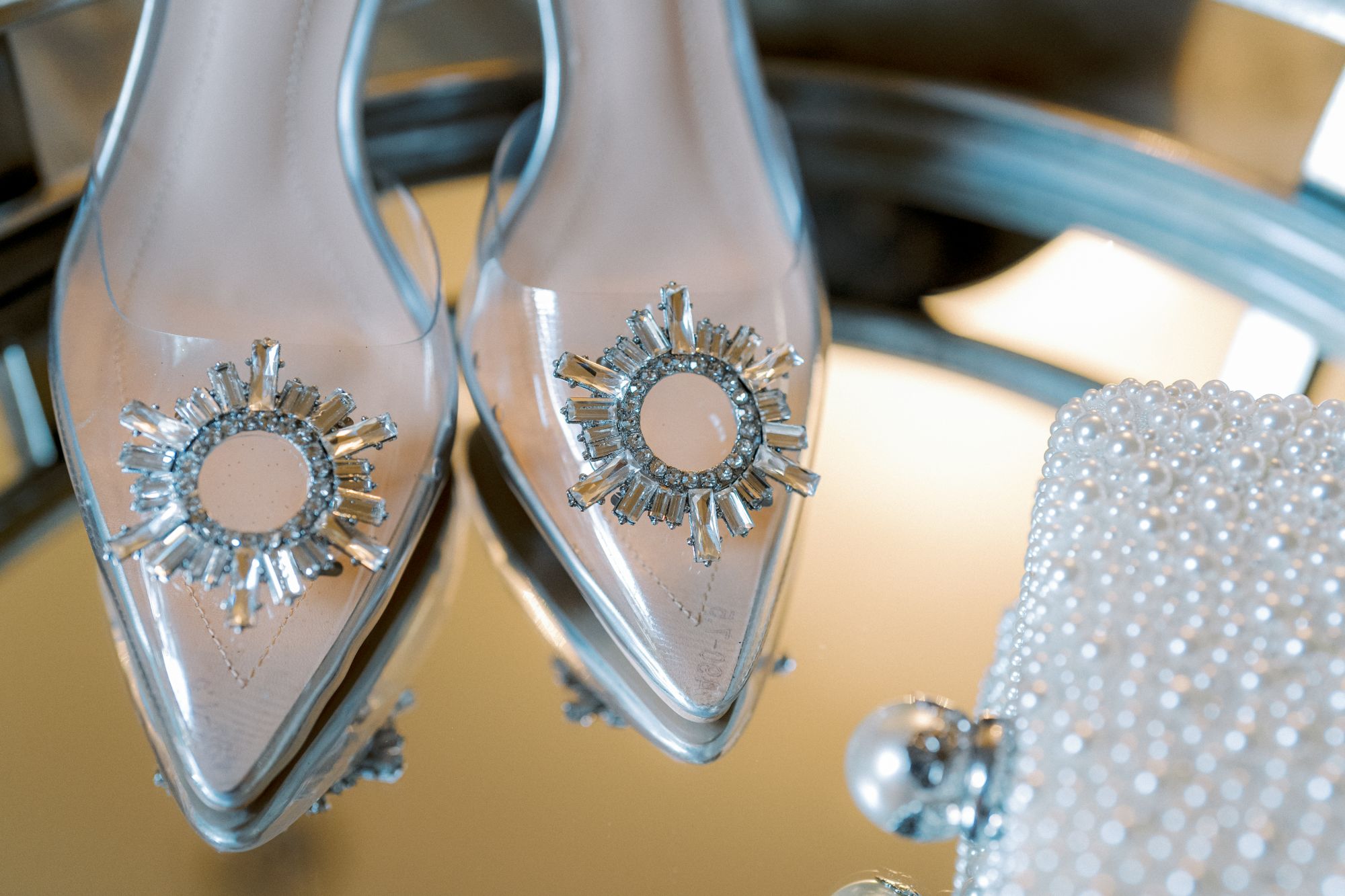 Close-up of elegant pointed-toe transparent heels with sparkling rhinestone embellishments, reflected on a mirror next to a pearl-studded clutch bag.