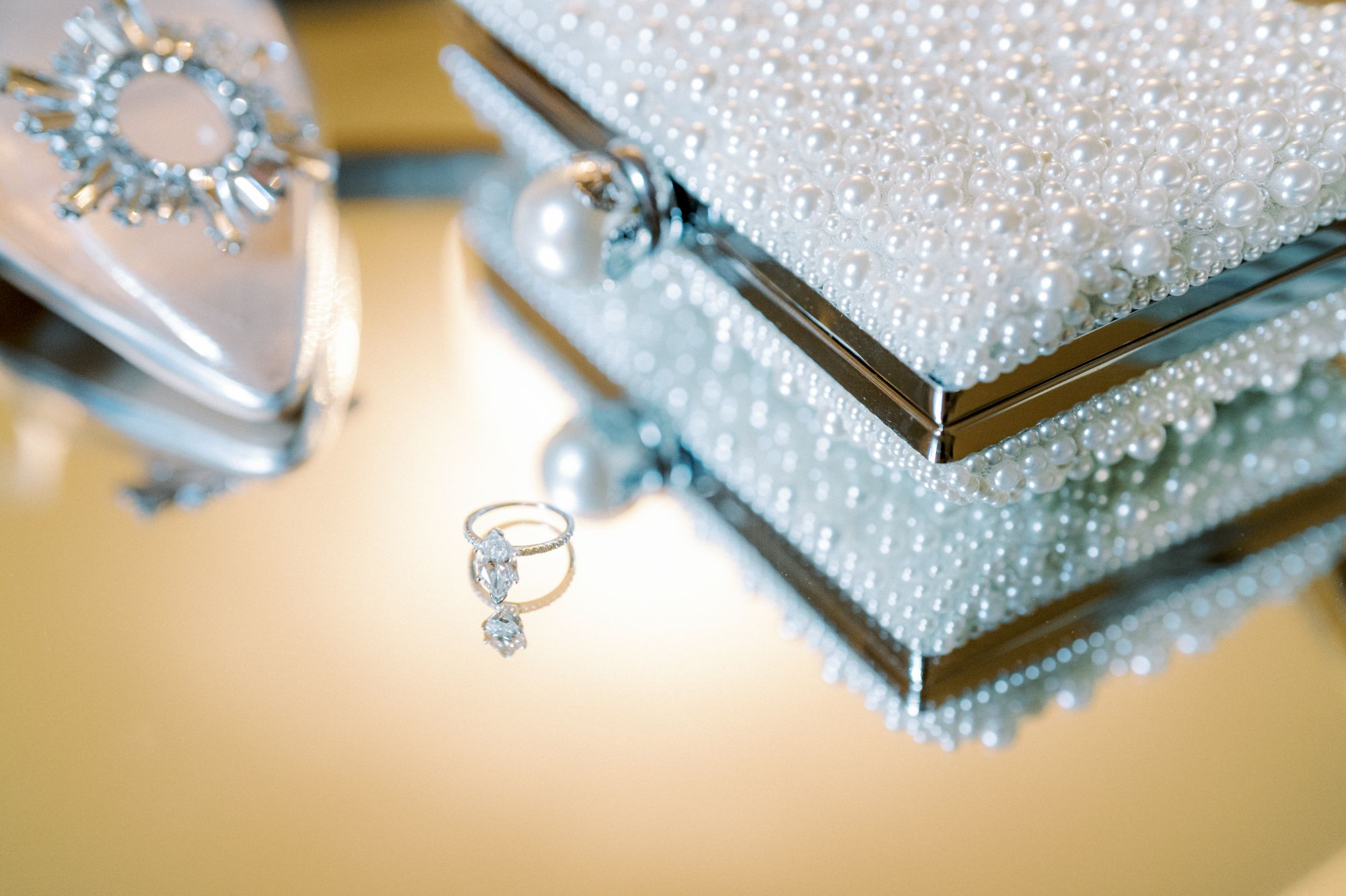 A diamond ring rests on a reflective surface beside a pearl-covered jewelry box, with part of a decorative shoe and a brooch visible in the background.