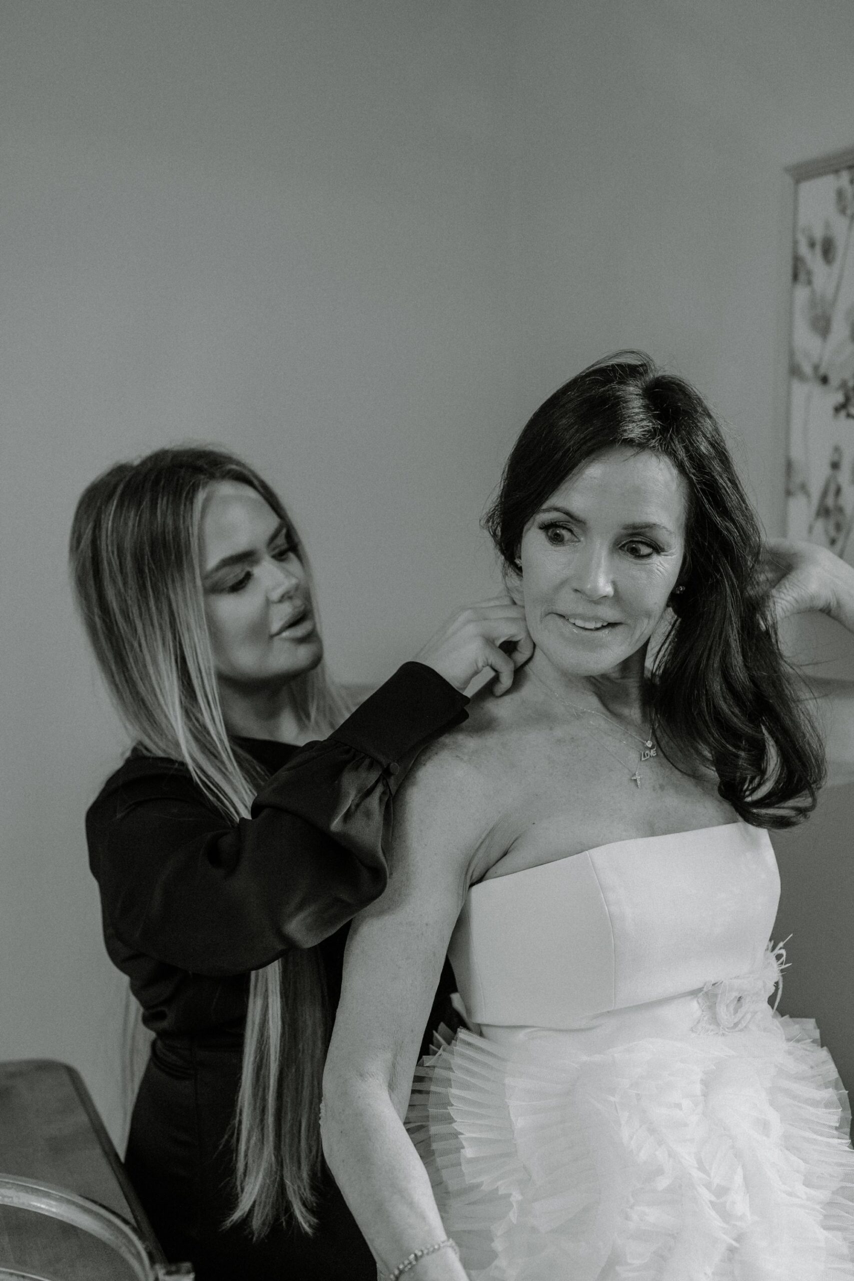 A woman in a white strapless dress is being helped with her necklace by another woman with long hair in a dark outfit. The scene appears calm and intimate, suggesting a special occasion like a wedding.