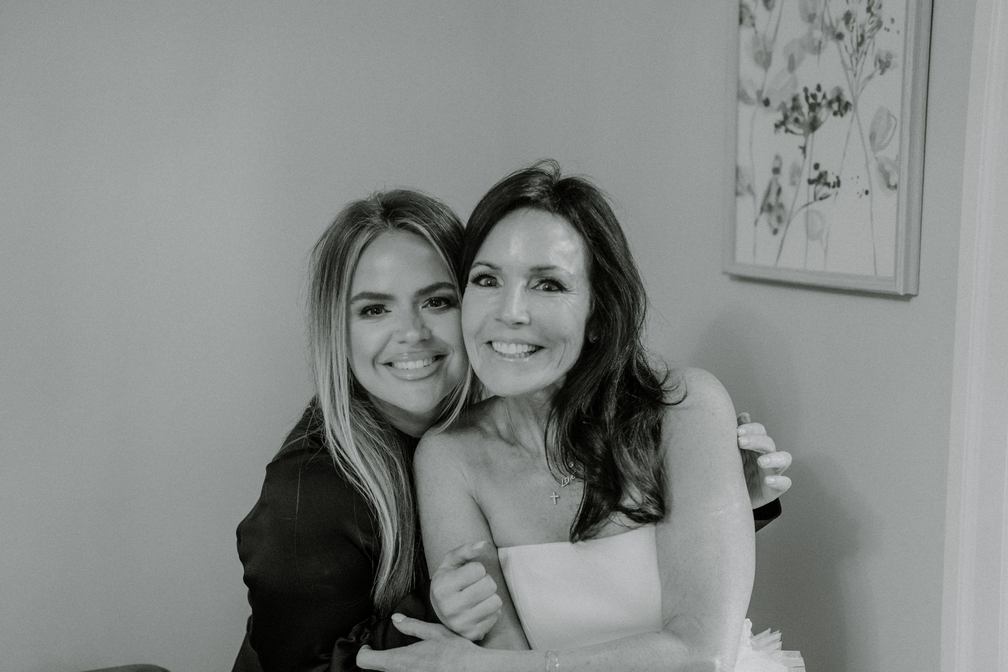 Two women smiling and hugging each other in a warmly lit room. One has long blonde hair and wears dark clothing, while the other has long brown hair and is dressed in white. A floral artwork hangs on the wall behind them.