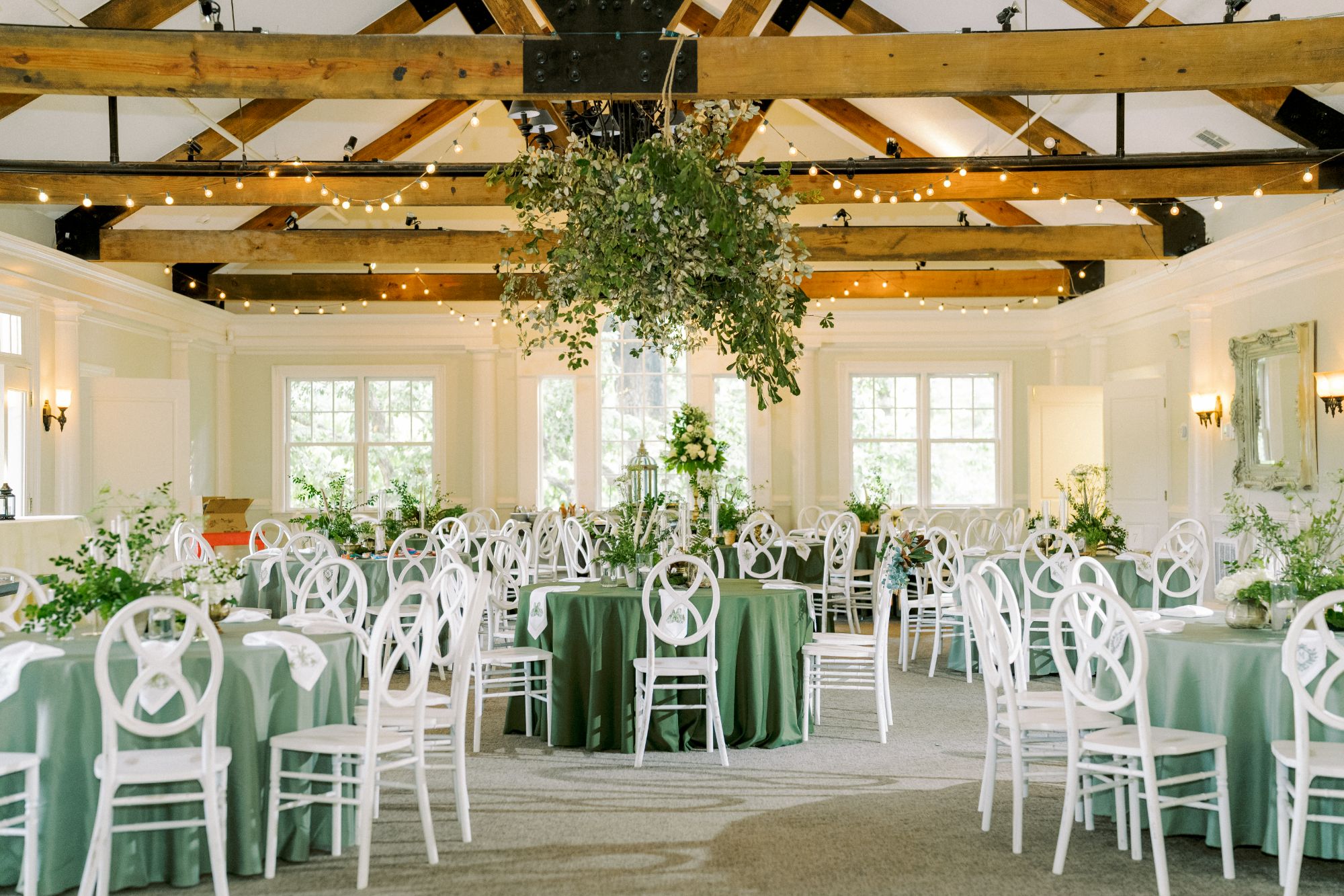 Elegant banquet hall with white chairs around green-clothed tables, greenery centerpieces, and a large hanging foliage arrangement. The room has wooden ceiling beams, string lights, and large windows letting in natural light.