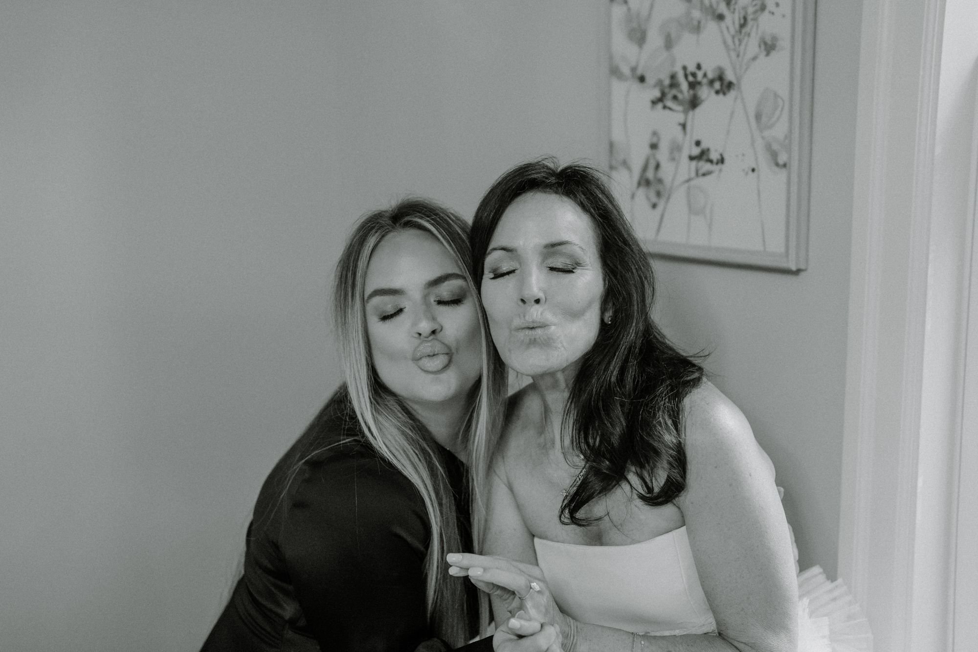 Two women pose playfully indoors, both facing the camera with their eyes closed and lips puckered as if blowing kisses. One wears a dark outfit, the other is in a light strapless dress. A floral painting hangs on the wall behind them.