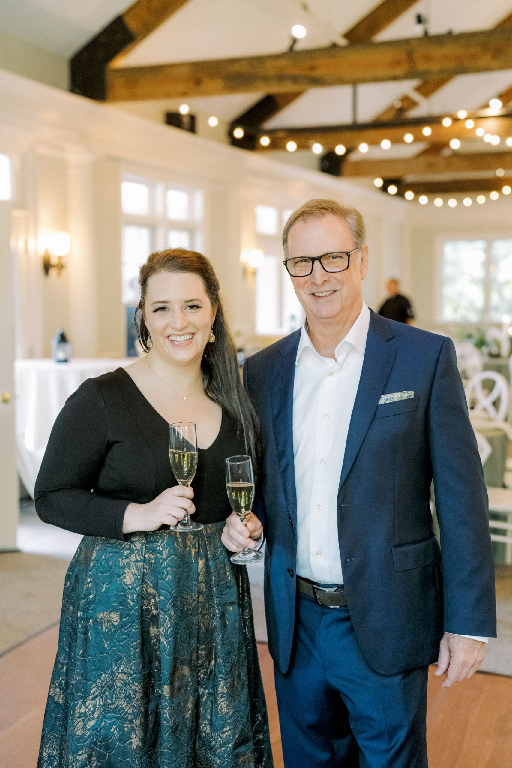 A smiling woman in a dark top and patterned skirt stands beside a man in a blue suit. They both hold glasses of champagne in a well-lit, elegant room with string lights and exposed wooden beams.