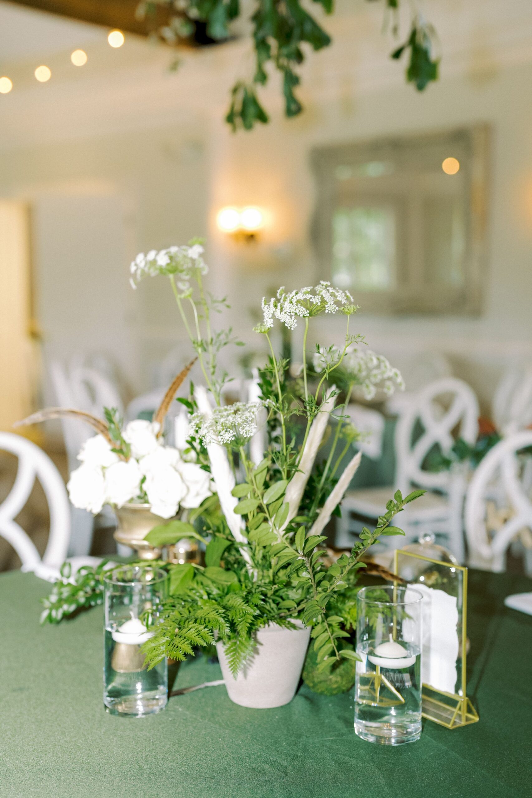 A green tablecloth with a floral centerpiece of white flowers, ferns, and greenery in a small pot, flanked by two glasses of water with lemon slices, in an elegant, softly lit room with white chairs.