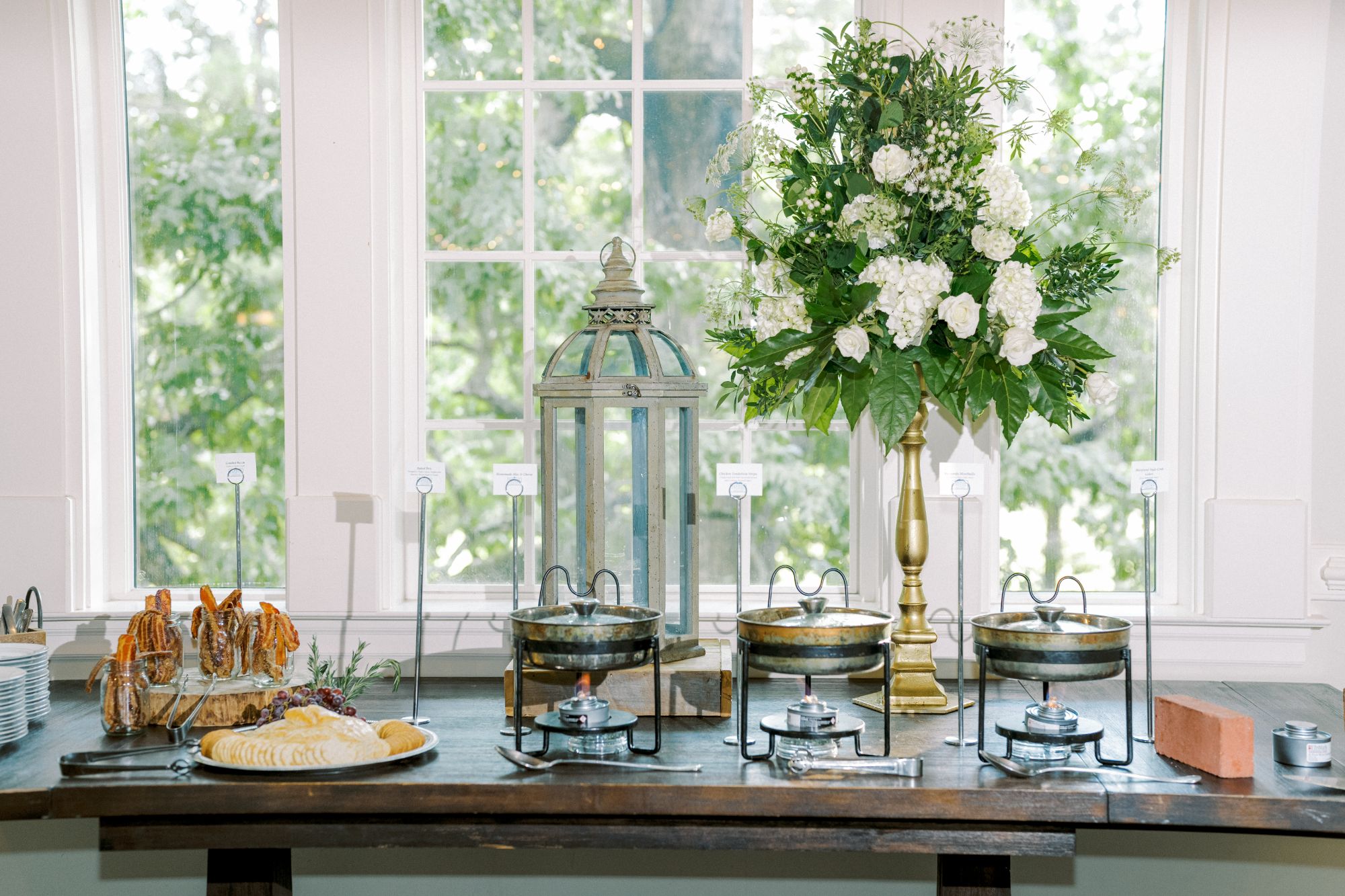 A buffet table set with chafing dishes, a cheese and pastry platter, plates, and tongs. A large floral arrangement and decorative lantern sit in front of bright windows with greenery visible outside.
