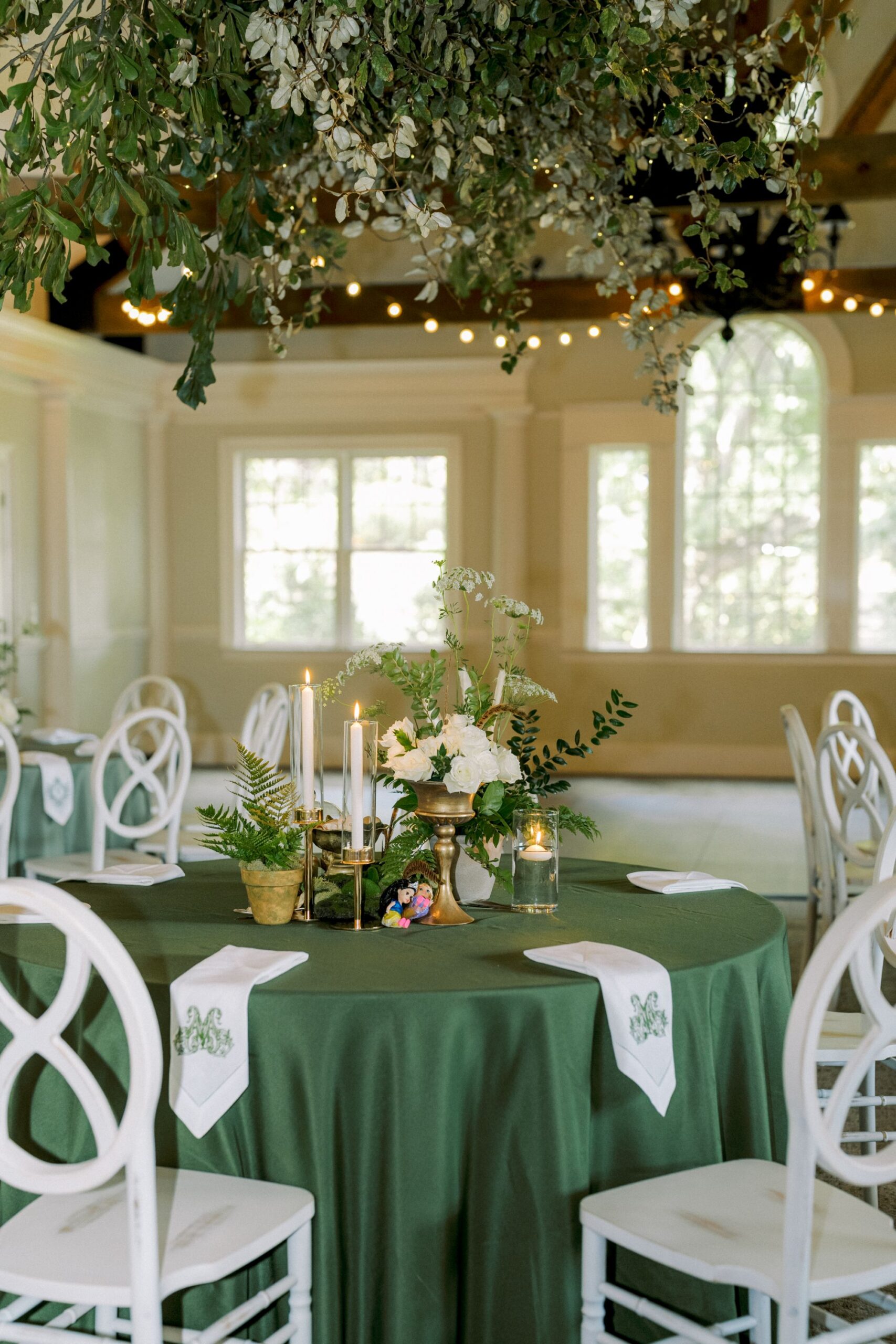 A round table with a green tablecloth is set with white napkins, candles, flowers, and greenery. White chairs surround the table, and leafy decorations hang above in a bright, elegant room with large windows.