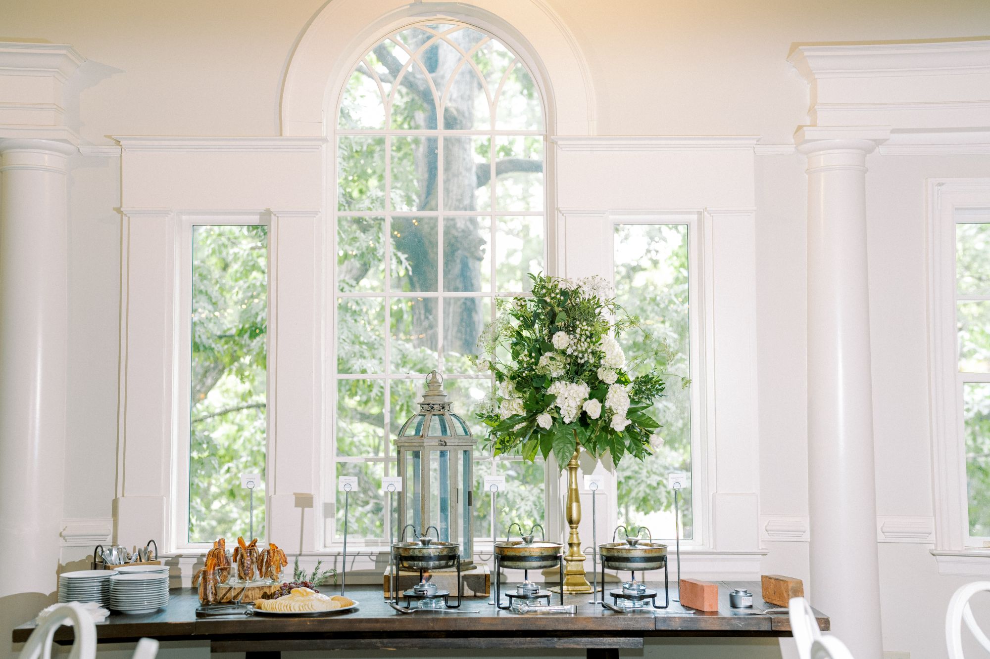A buffet table is set with plates, silverware, chafing dishes, and a cheese platter. A large bouquet of white flowers and greenery stands beside a lantern in front of a tall arched window with trees visible outside.