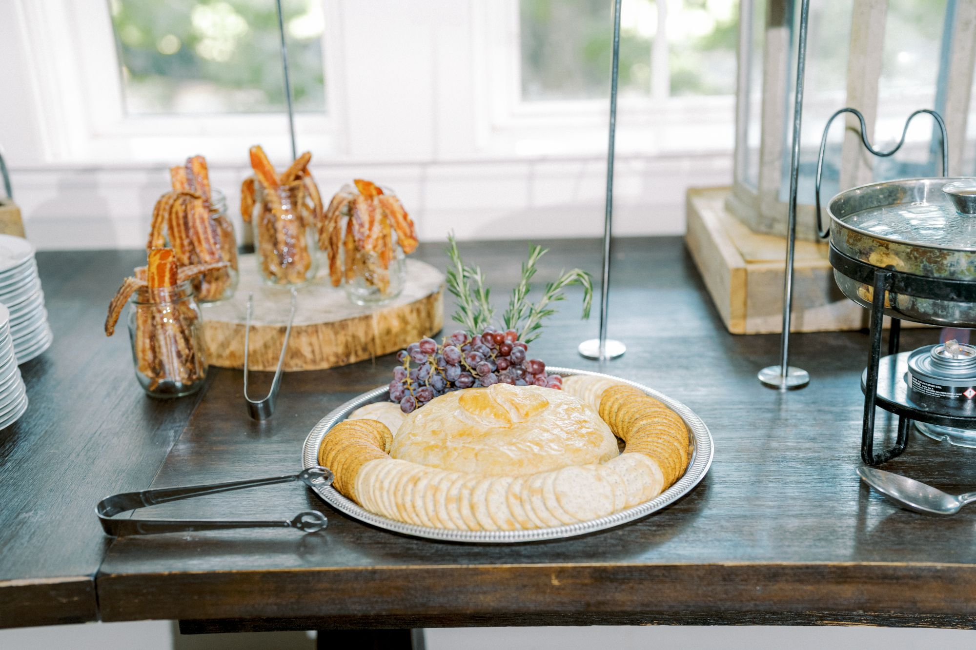 A platter of crackers and cheese spread garnished with grapes and herbs sits on a dark wooden table, with jars of pretzel sticks and serving tongs nearby. Light streams in from windows in the background.