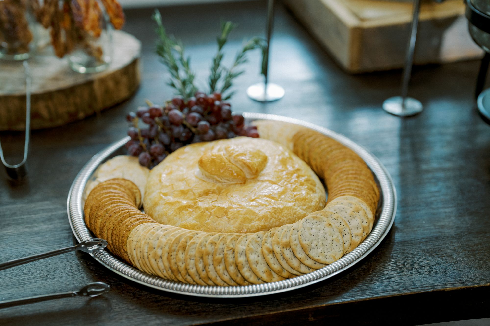 A round baked cheese wheel, topped with a decorative pastry, sits on a silver tray surrounded by assorted crackers and garnished with red grapes and rosemary. Metal tongs are placed at the edge of the tray.