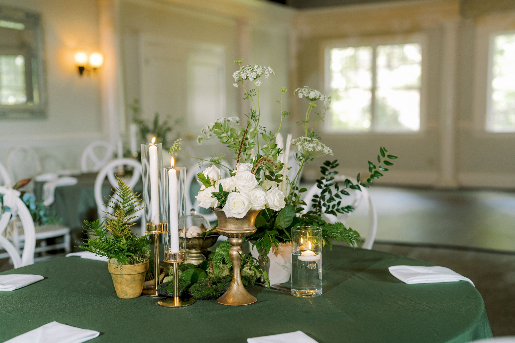 A round table with a green tablecloth is decorated with white napkins, gold candle holders, white flowers, greenery, and tall white candles in an elegant, sunlit room.