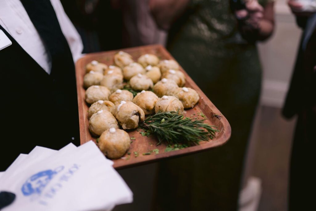 A server holds a wooden tray of bite-sized pastry appetizers garnished with herbs, with a stack of napkins on the side. People in formal attire are seen in the blurred background.