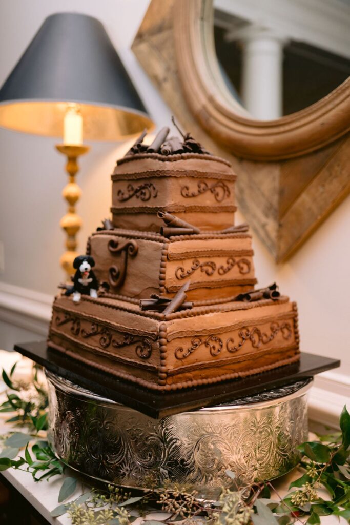 A three-tiered chocolate cake with ornate piping and chocolate decorations sits on a silver stand. A small figure of a dog decorates one corner. The cake is displayed on a table with greenery, next to a lamp and mirror.