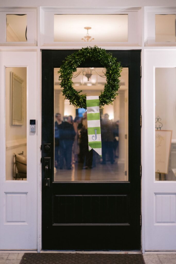 A green wreath with a green and white striped ribbon hangs on a black front door, which has glass panes showing a group of people gathered inside a brightly lit entryway.